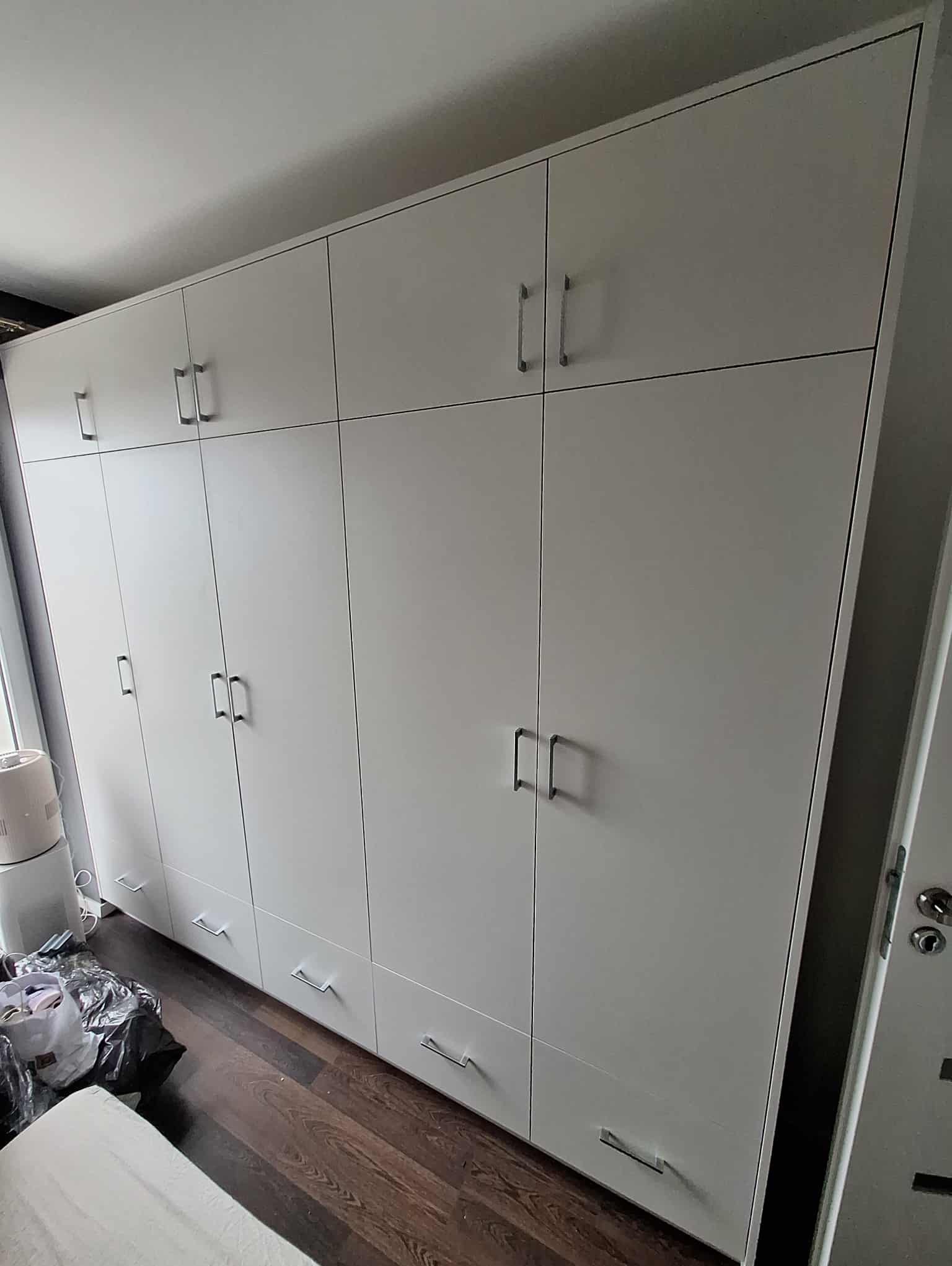 Duża, biała szafa wnękowa z szufladami i metalowymi uchwytami, umieszczona w pokoju z drewnianą podłogą. Widok z perspektywy lekko od dołu.