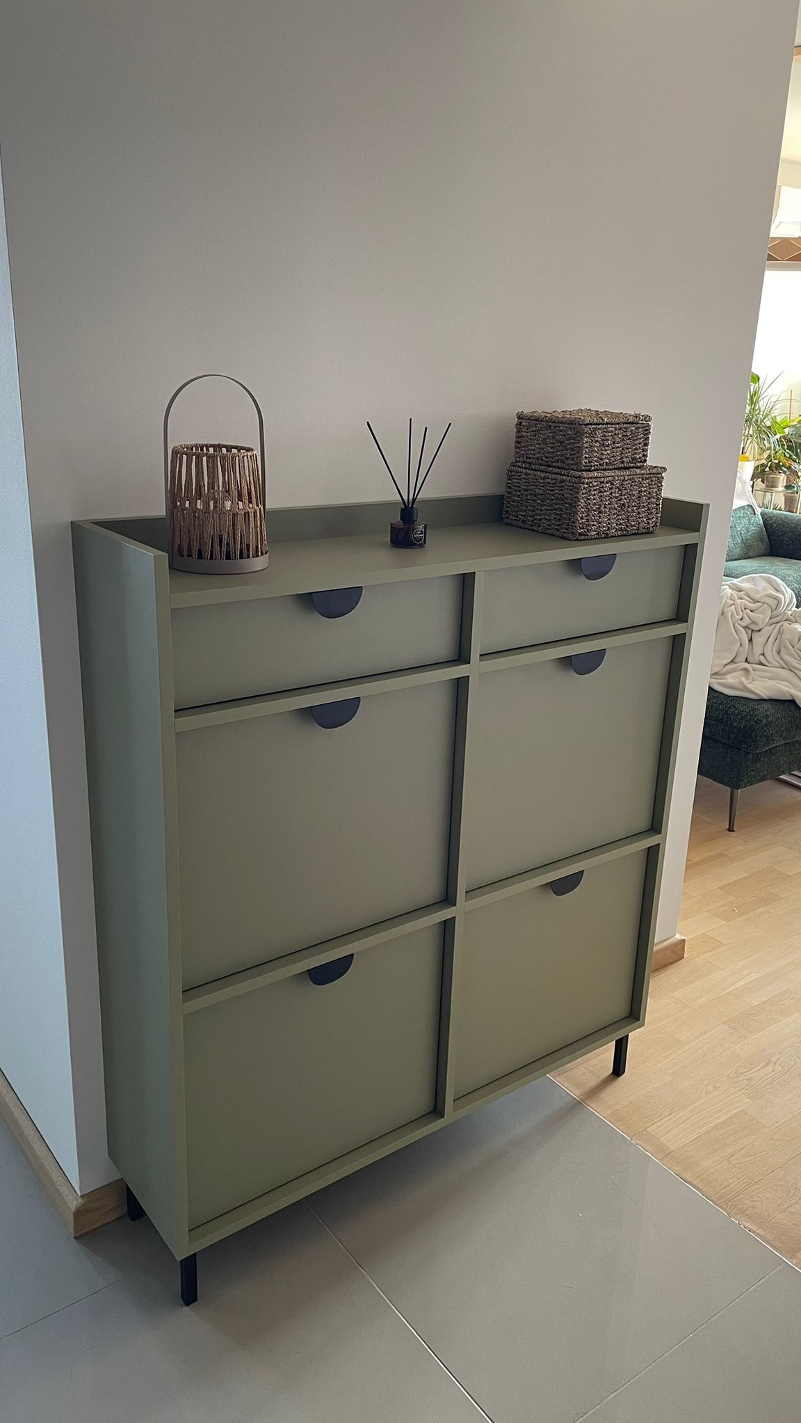 Stylowa, oliwkowa szafka na buty z sześcioma szufladami, z czarnymi uchwytami, udekorowana lampionem i dyfuzorem zapachowym, na tle jasnego wnętrza.