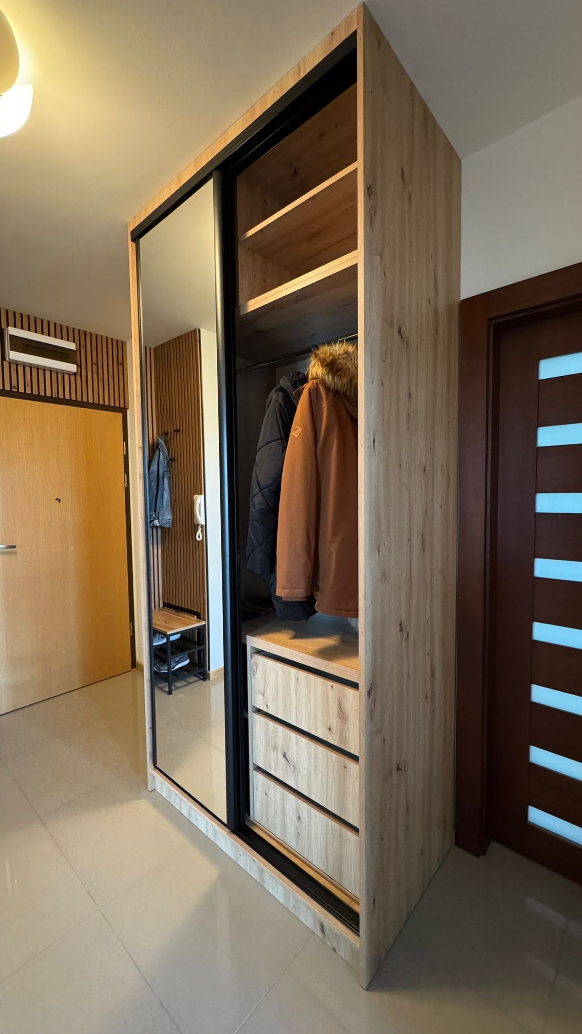 Szafa wnękowa z lustrem i drewnianym wykończeniem, z widocznymi ubraniami wewnątrz. Elementy wystroju wnętrza w tle, w tym drzwi i panele ścienne.