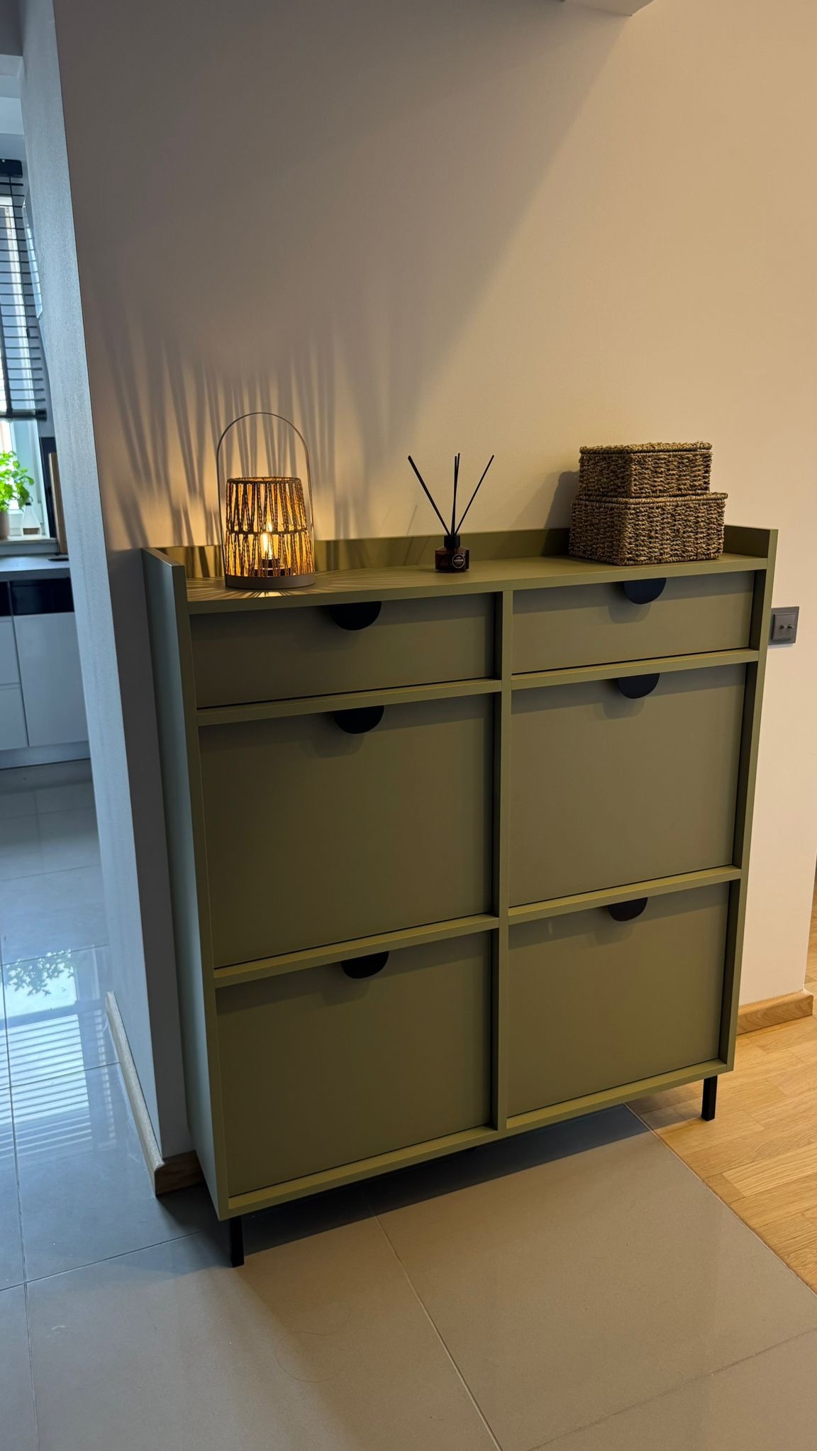 Funkcjonalna szafka na buty w odcieniach zieleni z minimalistycznymi uchwytami, uzupełniona o dekoracyjną lampę i koszyki z wikliny w nowoczesnym wnętrzu.