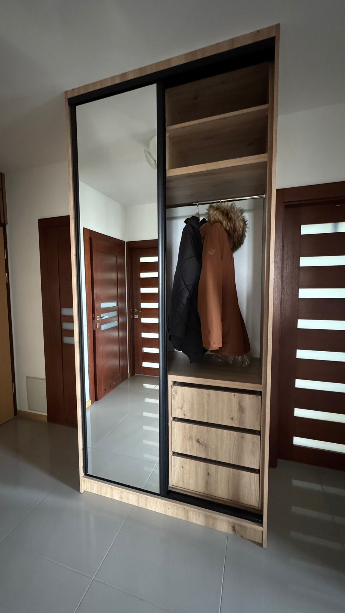 Szafa wnękowa z przesuwnymi drzwiami lustrzanymi i drewnianymi szufladami, widoczne ubrania w środku. Ujęcie z niskiego kąta.