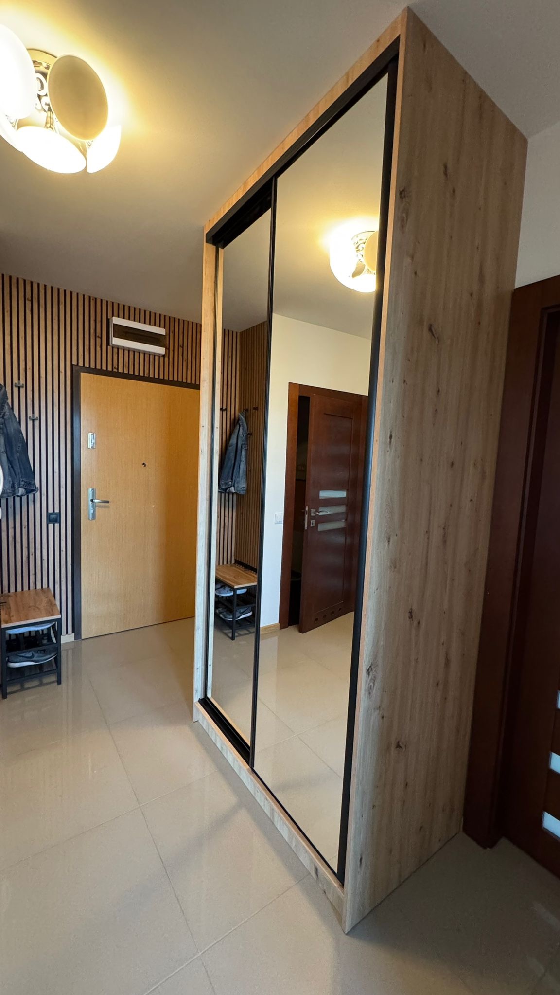 Szafa wnękowa z lustrzanymi drzwiami przesuwnymi, wykończona drewnopodobnym dekorem, na tle przedpokoju z panelami ściennymi i jasną podłogą z płytek.