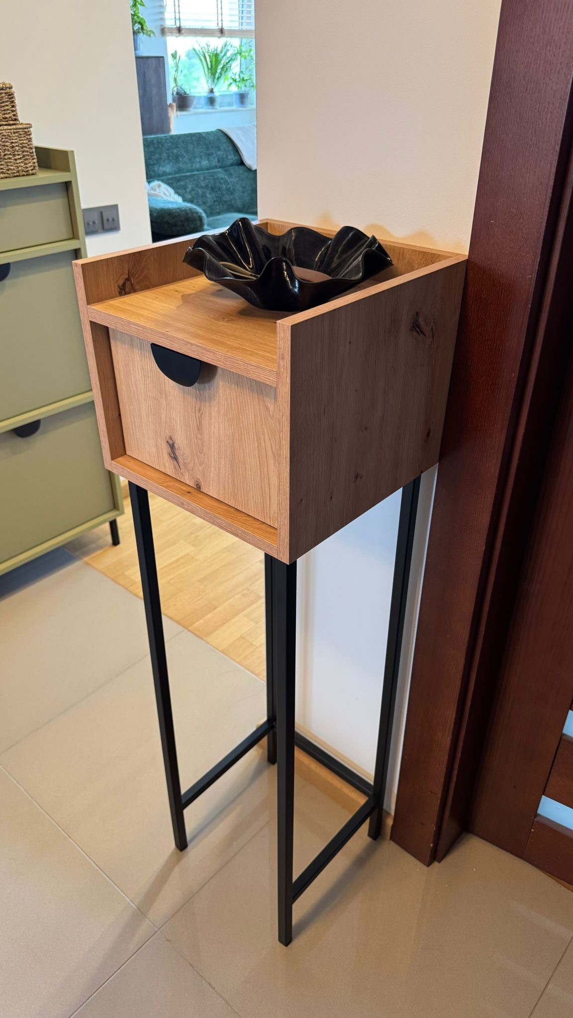 Drewniana szafka nocna na metalowych nogach z czarną misą dekoracyjną. Widoczny fragment drzwi i innej szafki w tle. Jasne wnętrze.