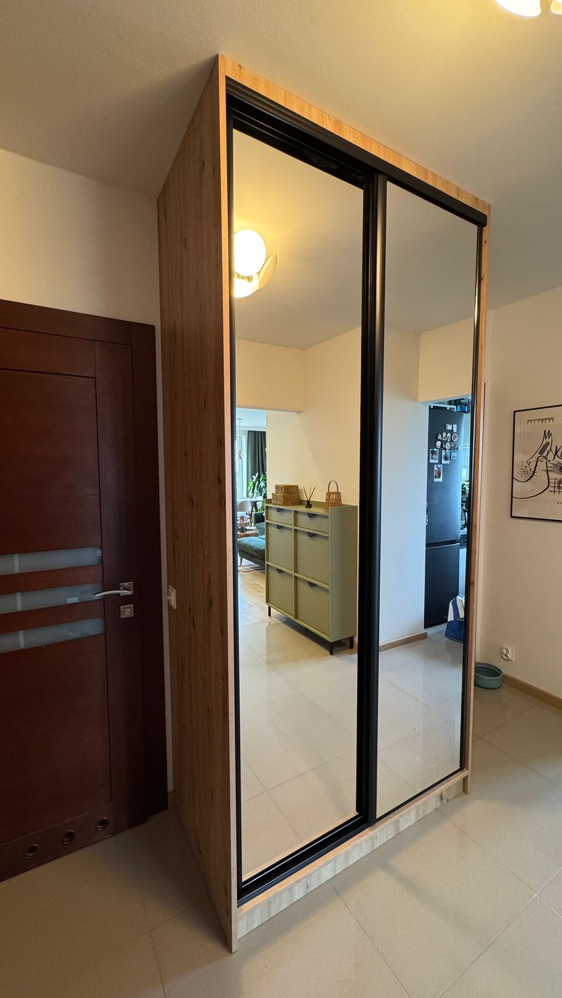 Szafa wnękowa z lustrzanymi drzwiami przesuwnymi, drewniane wykończenie, czarne ramy. Widok na wnętrze mieszkania przez lustro.