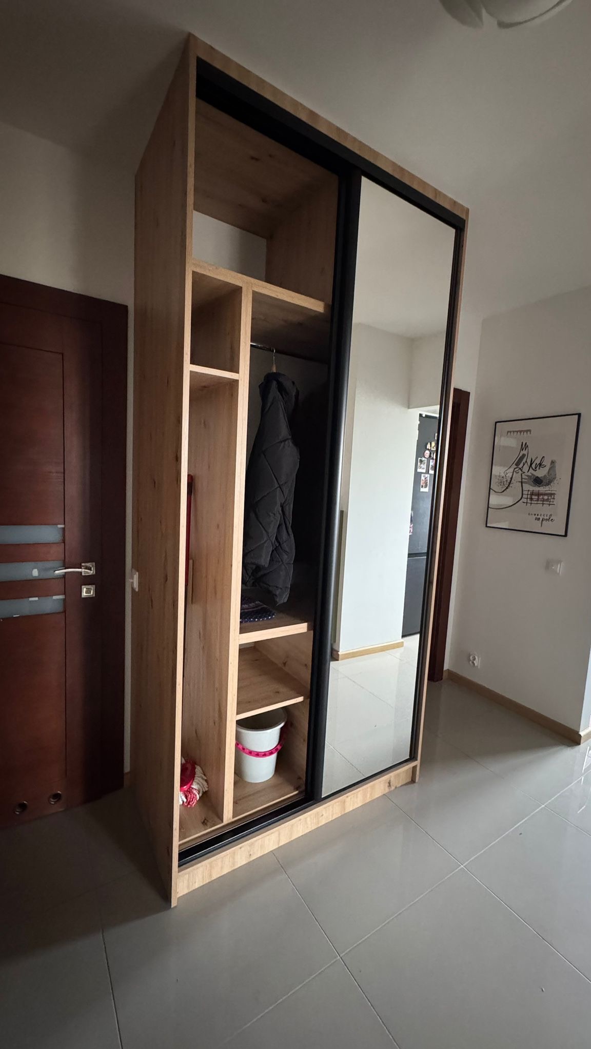 Jasna szafa z lustrem i czarnymi detalami, z otwartymi półkami. Widoczna kurtka w środku. Ujęcie z perspektywy podłogi.