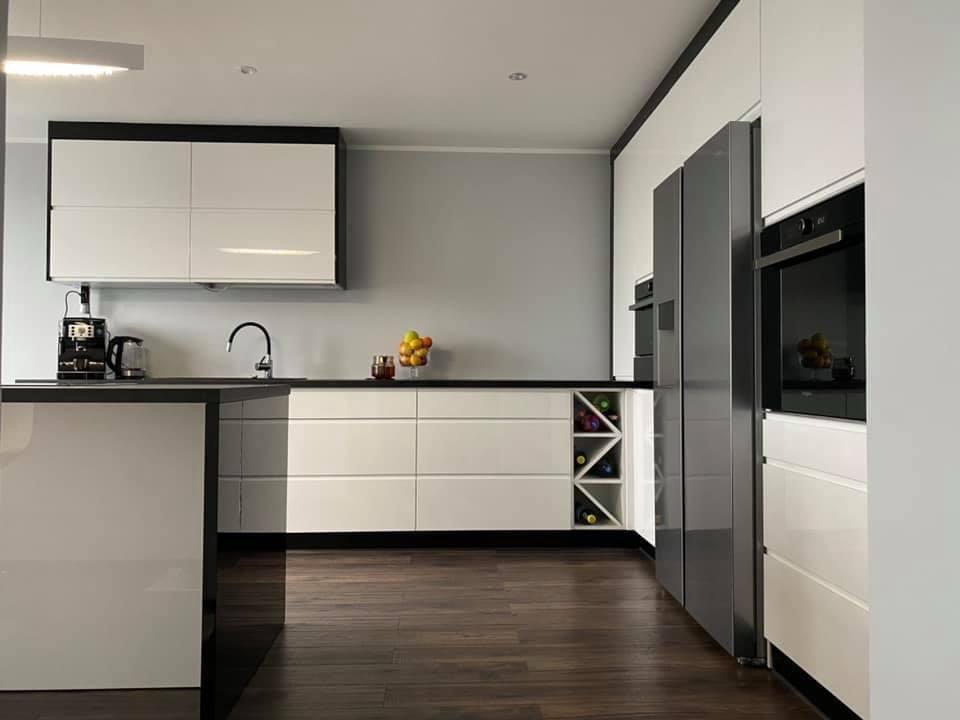 Nowoczesna kuchnia z białymi, lakierowanymi frontami, czarnymi blatami i akcentami. Widoczny regał na wino i sprzęt AGD w zabudowie. Parkiet na podłodze.