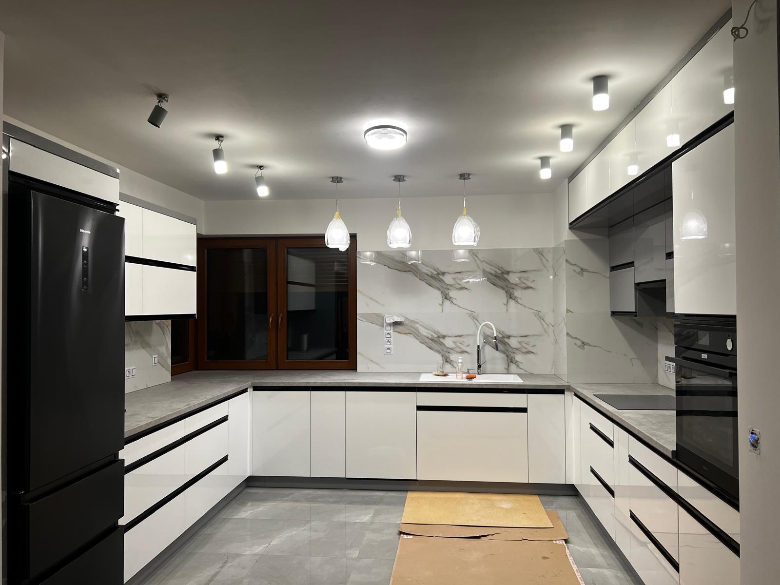 Nowoczesna kuchnia w bieli i czerni z marmurowym backsplashiem, oświetleniem punktowym i wiszącymi lampami. Minimalistyczny design z elementami dekoracyjnymi.