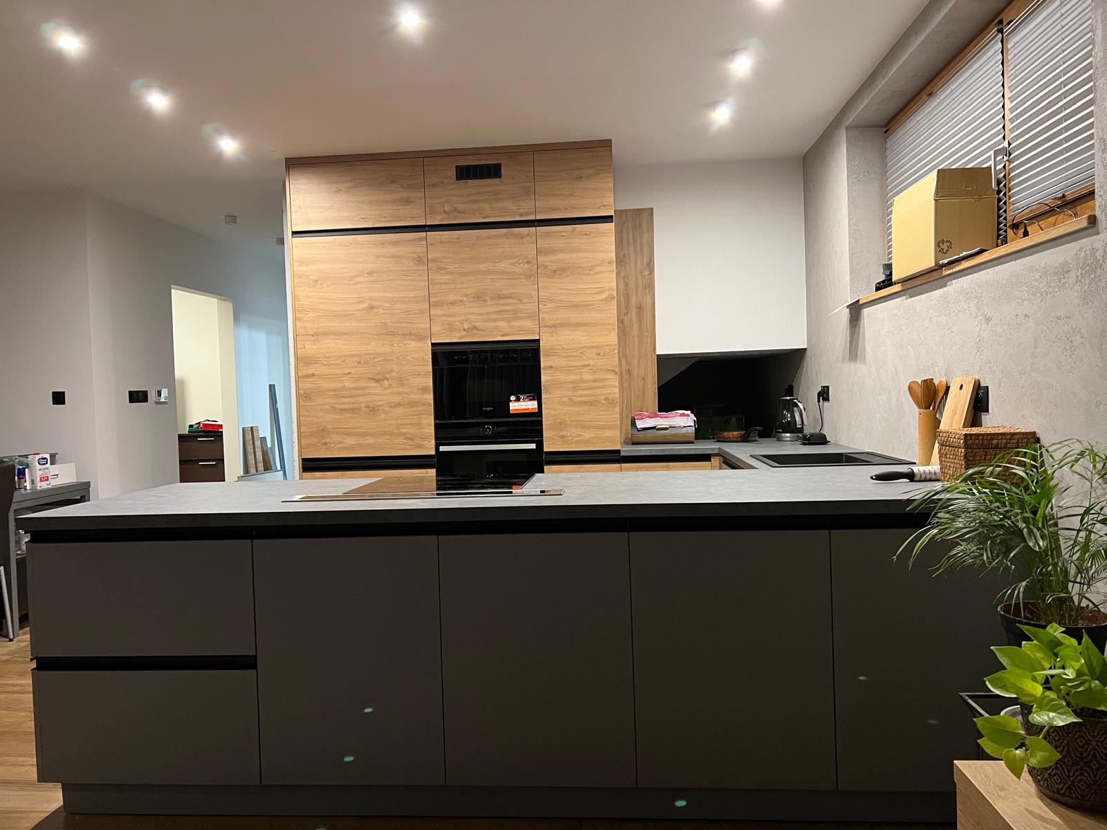 Nowoczesna kuchnia z wyspą i zabudową drewnopodobną z czarnymi akcentami, oświetleniem punktowym i roślinami doniczkowymi na blacie.