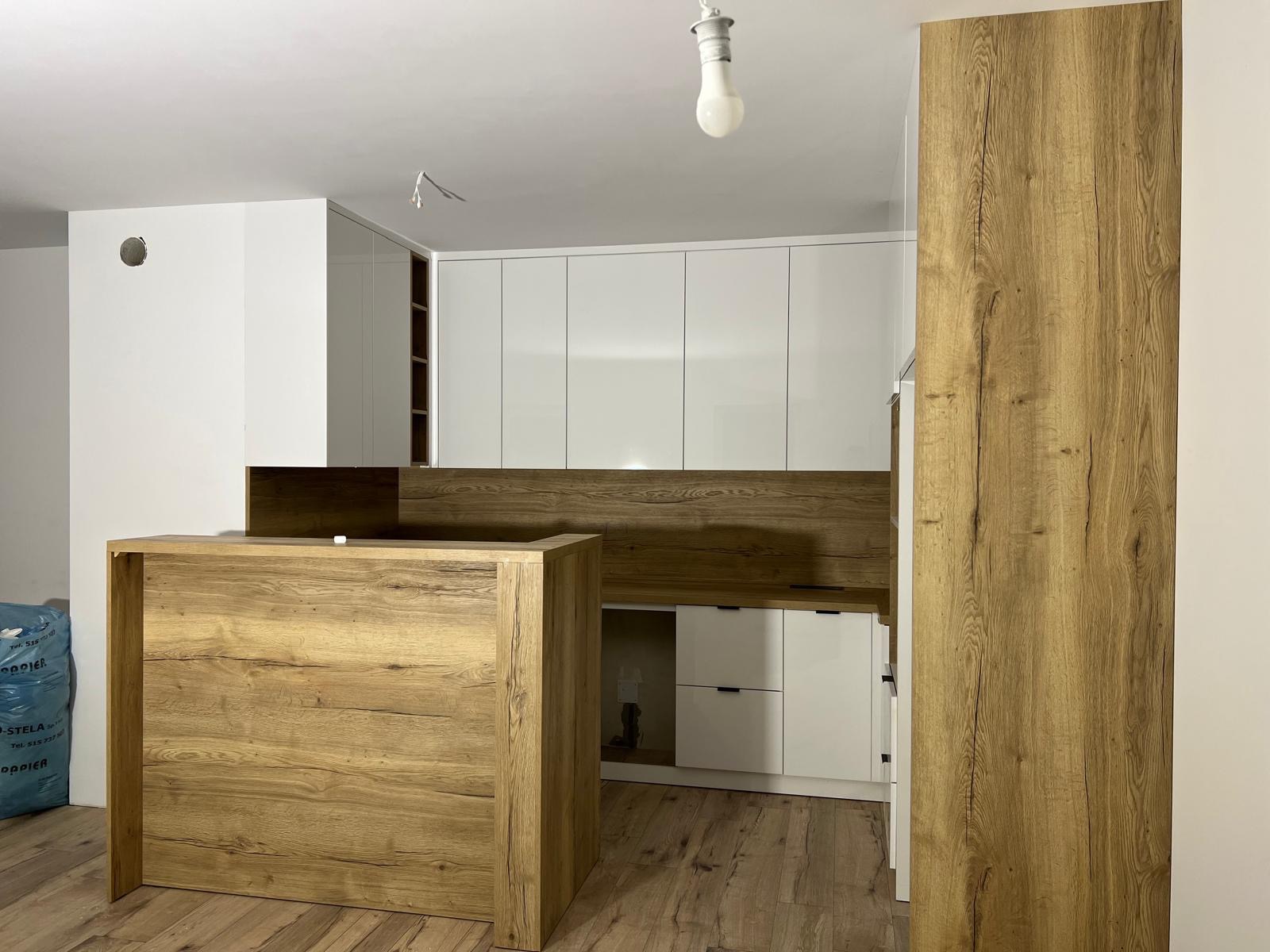 Nowoczesna kuchnia z białą zabudową i drewnianym blatem. Widoczny barek z drewnianym wykończeniem i podłoga imitująca drewno. W tle torba budowlana.