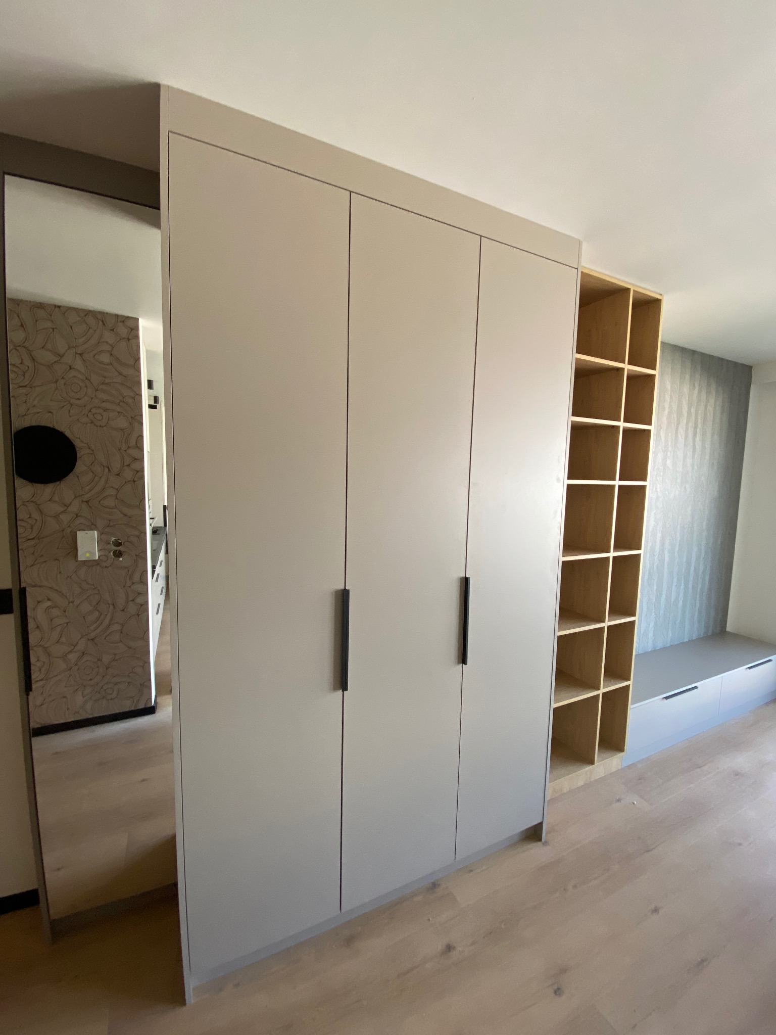 Jasnoszara szafa z trzema drzwiami i czarnymi uchwytami, z lustrem po lewej stronie i regałem z półkami po prawej, na tle jasnego parkietu.