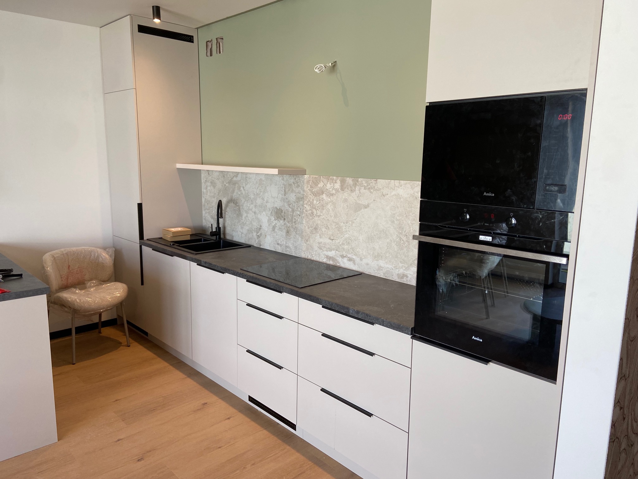Nowoczesna kuchnia z białymi frontami, czarnym blatem i sprzętem Amica. Uwagę zwraca krzesło zabezpieczone folią. Ściana w odcieniach zieleni i marmuru.