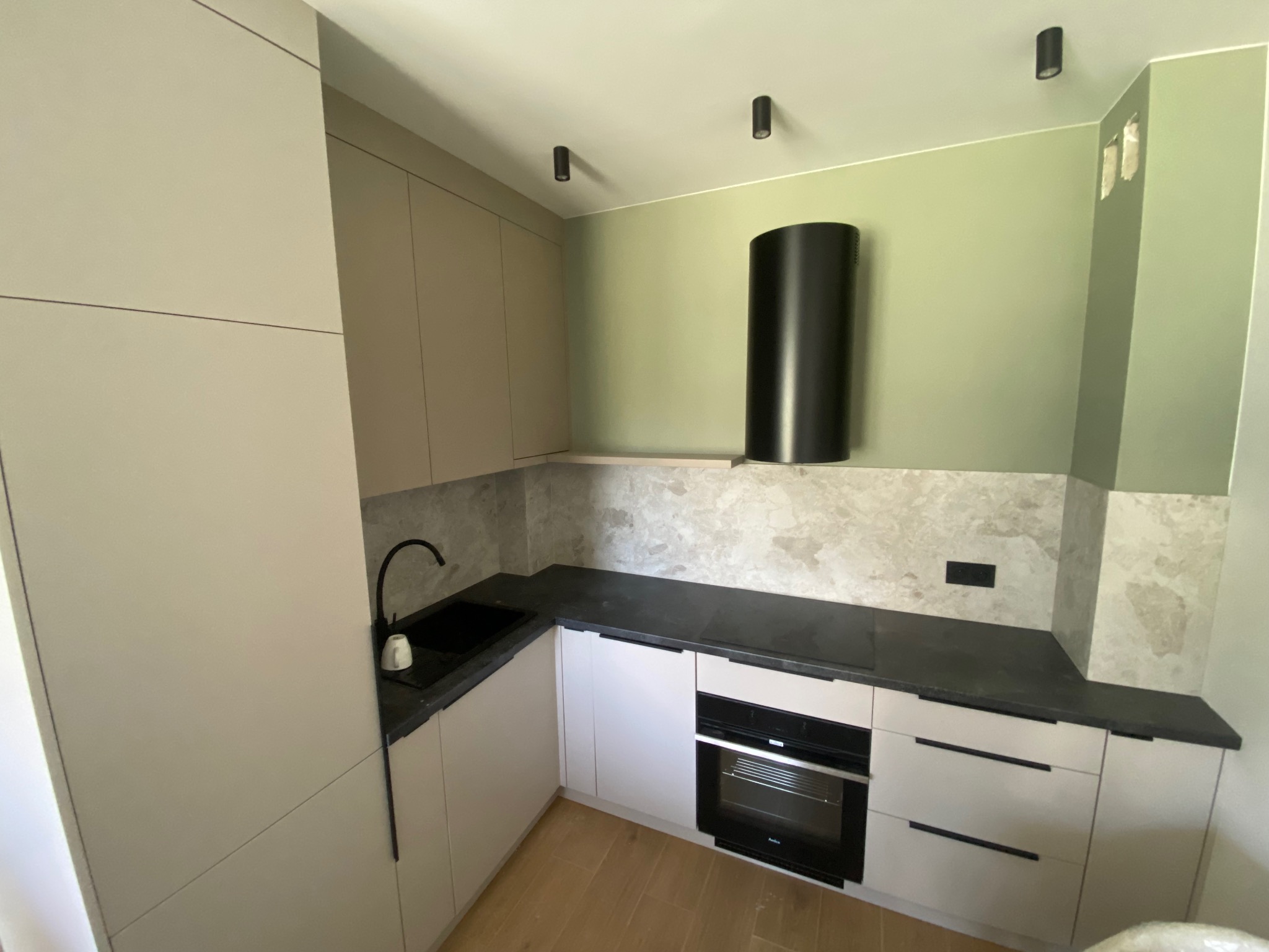 Jasna, nowoczesna kuchnia z beżowymi szafkami, czarnym blatem i sprzętem AGD. Uwagę zwraca okrągły okap w kolorze czarnym. Ściana w odcieniach zieleni.