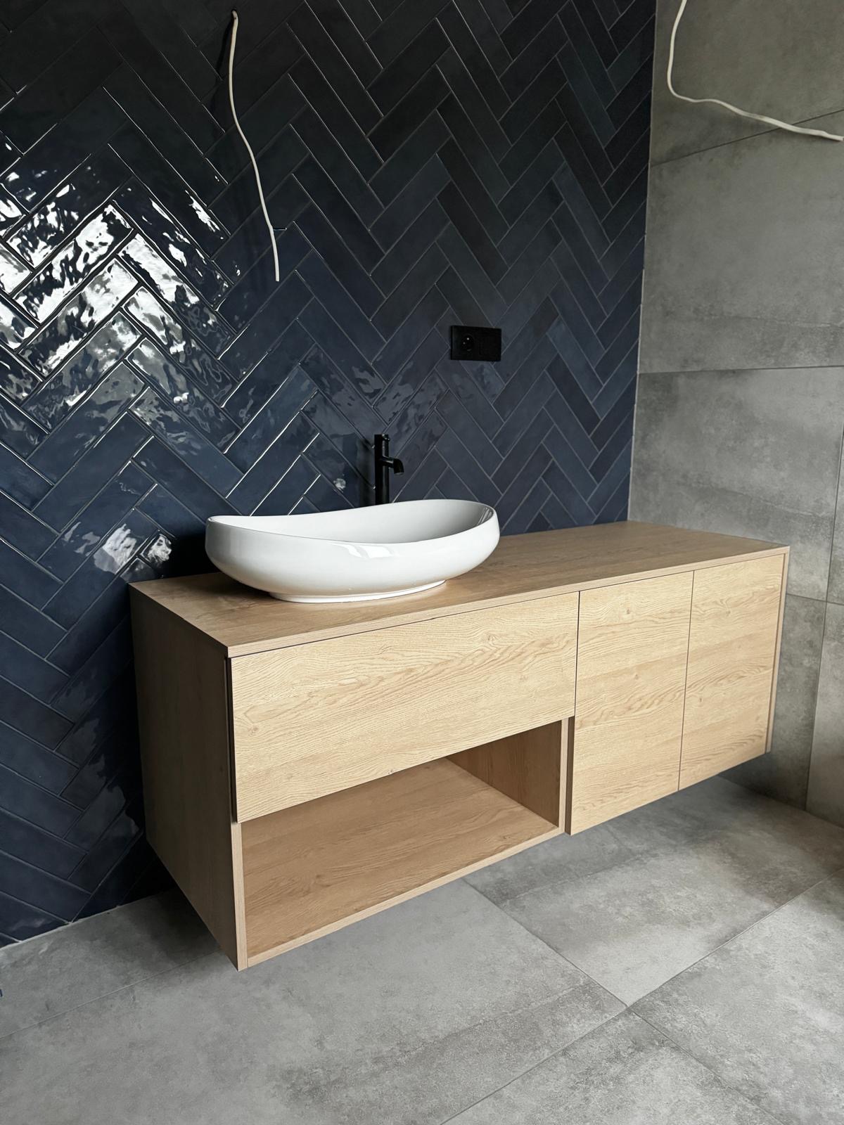 Nowoczesna łazienka: drewniana szafka z owalną umywalką na tle ciemnych, błyszczących płytek ułożonych w jodełkę. Minimalistyczny design, surowy beton na podłodze.