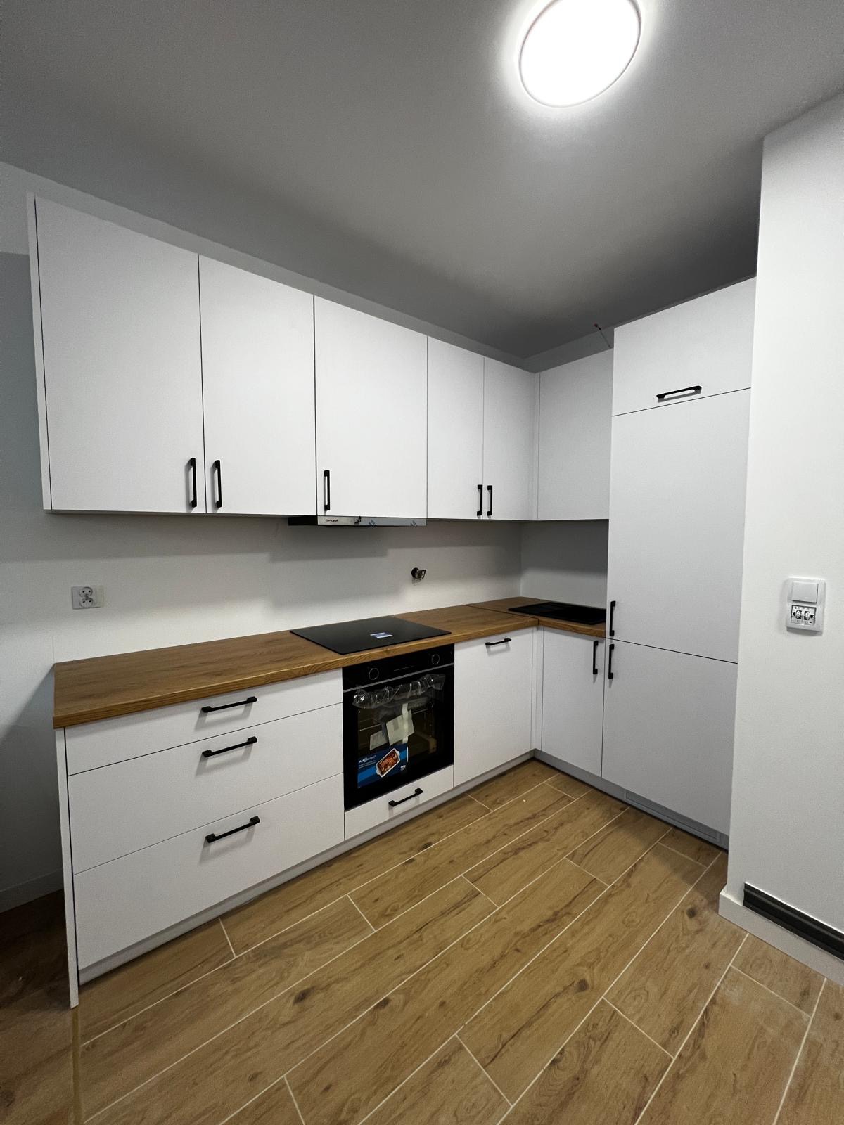 Nowoczesna, biała kuchnia z drewnianym blatem i czarnymi uchwytami, widok na zabudowę kuchenną z piekarnikiem i płytą grzewczą, oświetlenie sufitowe.