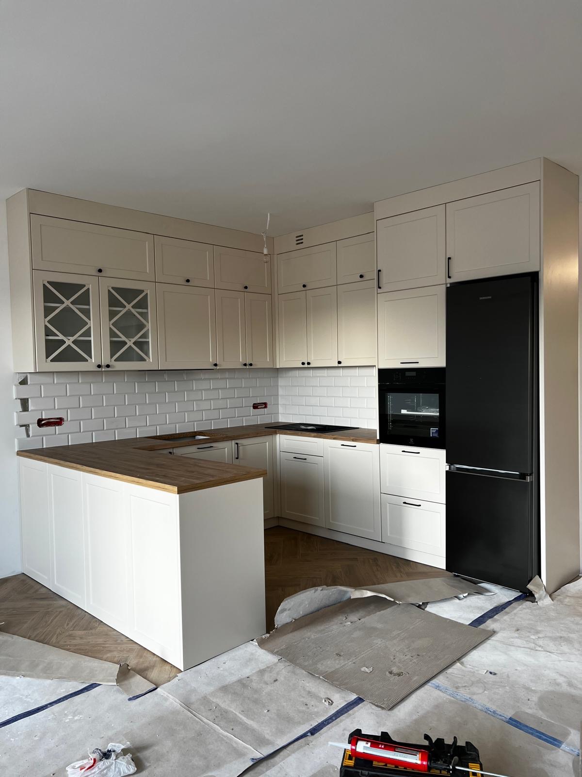 Jasna, nowoczesna kuchnia na wymiar z drewnianym blatem i czarną lodówką. Widoczne płytki na ścianie i fragment podłogi. Częściowo zabezpieczona podłoga.
