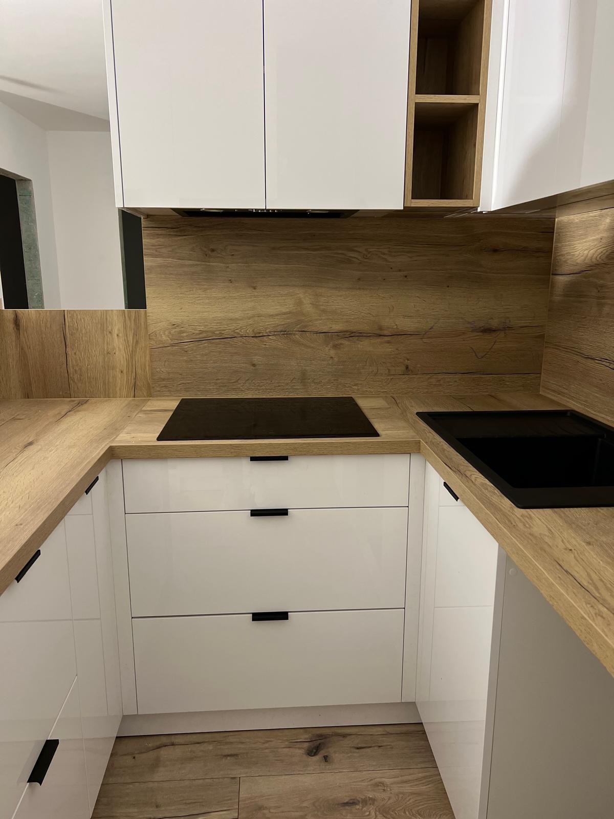 Nowoczesna kuchnia: białe, lakierowane fronty szafek z czarnymi uchwytami, drewniany blat i panel nad blatem, czarna płyta indukcyjna i zlewozmywak.