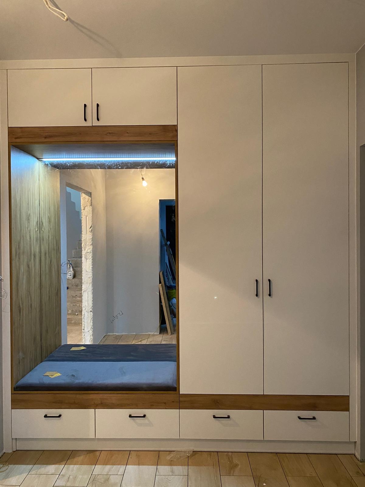 Biała szafa wnękowa z drewnianym akcentem i siedziskiem, oświetlona listwą LED. Widok na wnętrze przez otwartą przestrzeń. Minimalistyczny design.