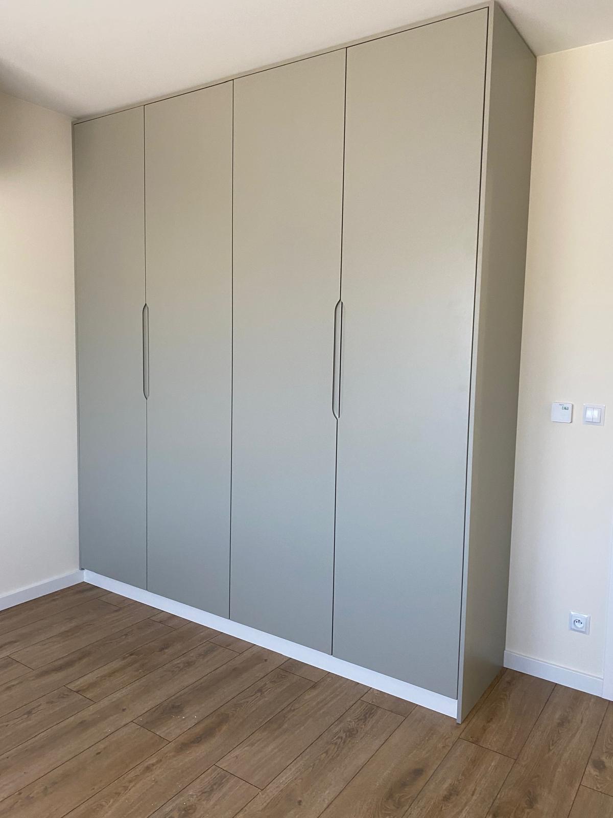Szara, minimalistyczna szafa wnękowa z czterema drzwiami, z podłużnymi uchwytami, dopasowana do jasnego wnętrza z drewnianą podłogą.