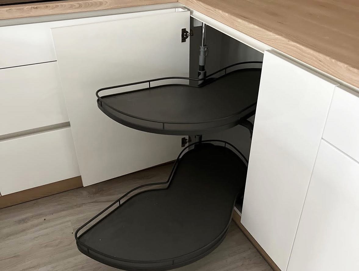 Wnętrze szafki narożnej z półkami obrotowymi w nowoczesnej kuchni. Białe fronty, drewniany blat i szare półki tworzą spójny design.