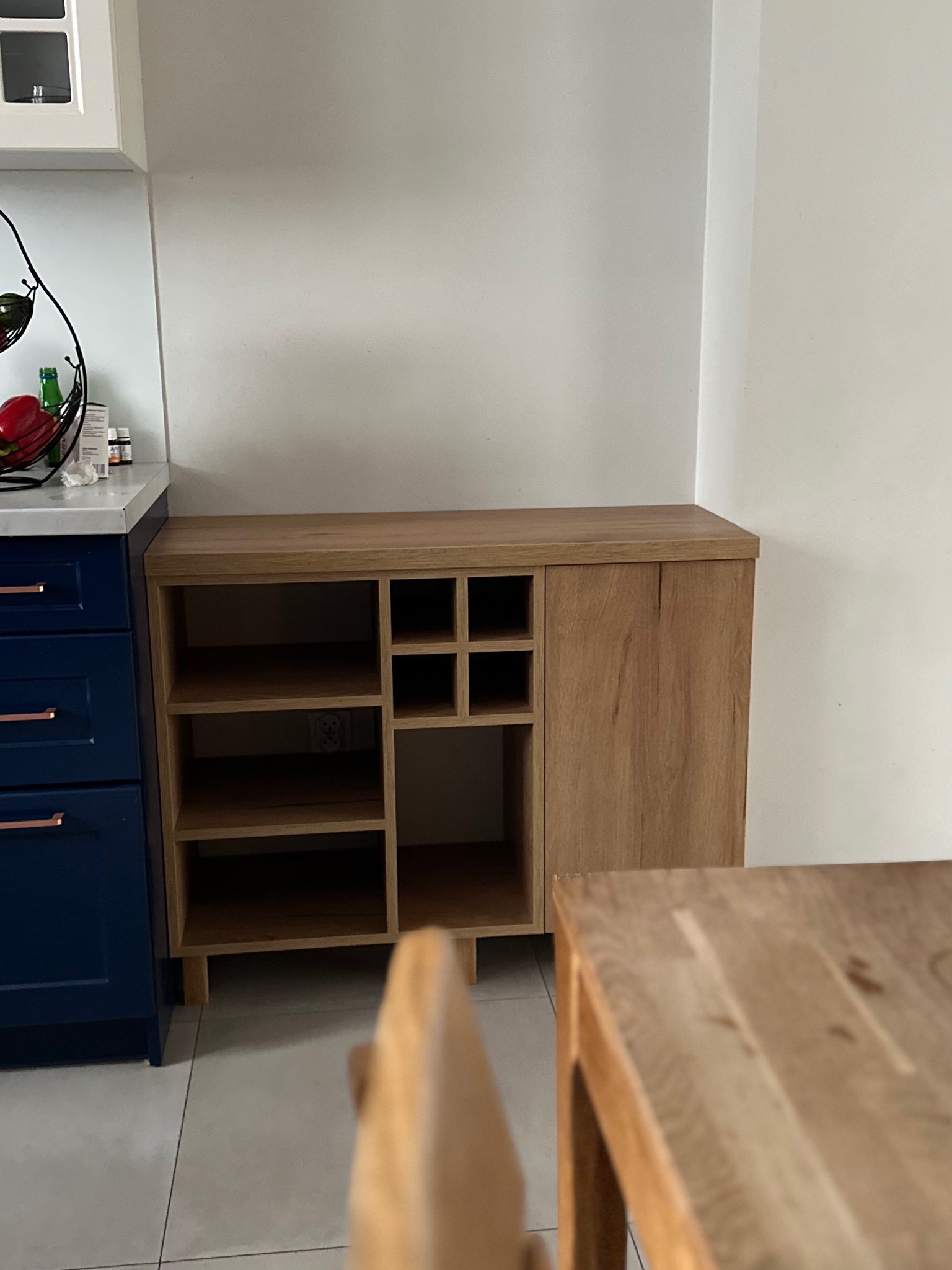 Drewniana szafka z otwartymi półkami i szafką z drzwiczkami, stojąca obok niebieskich szafek kuchennych. Widoczny fragment drewnianego stołu.