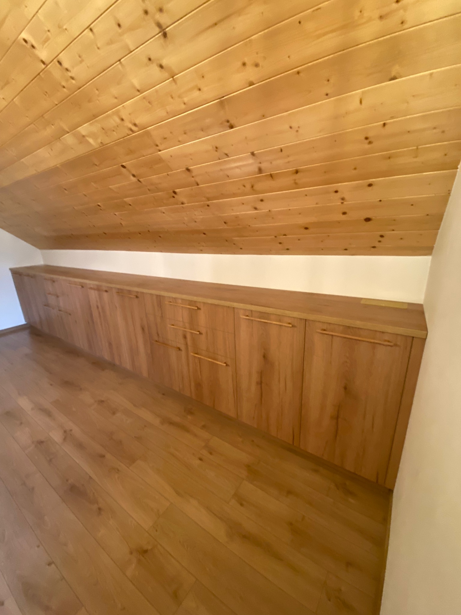 Drewniana szafa wnękowa z długimi, minimalistycznymi uchwytami, dopasowana do skosu poddasza. Podłoga i sufit również wykończone drewnem.