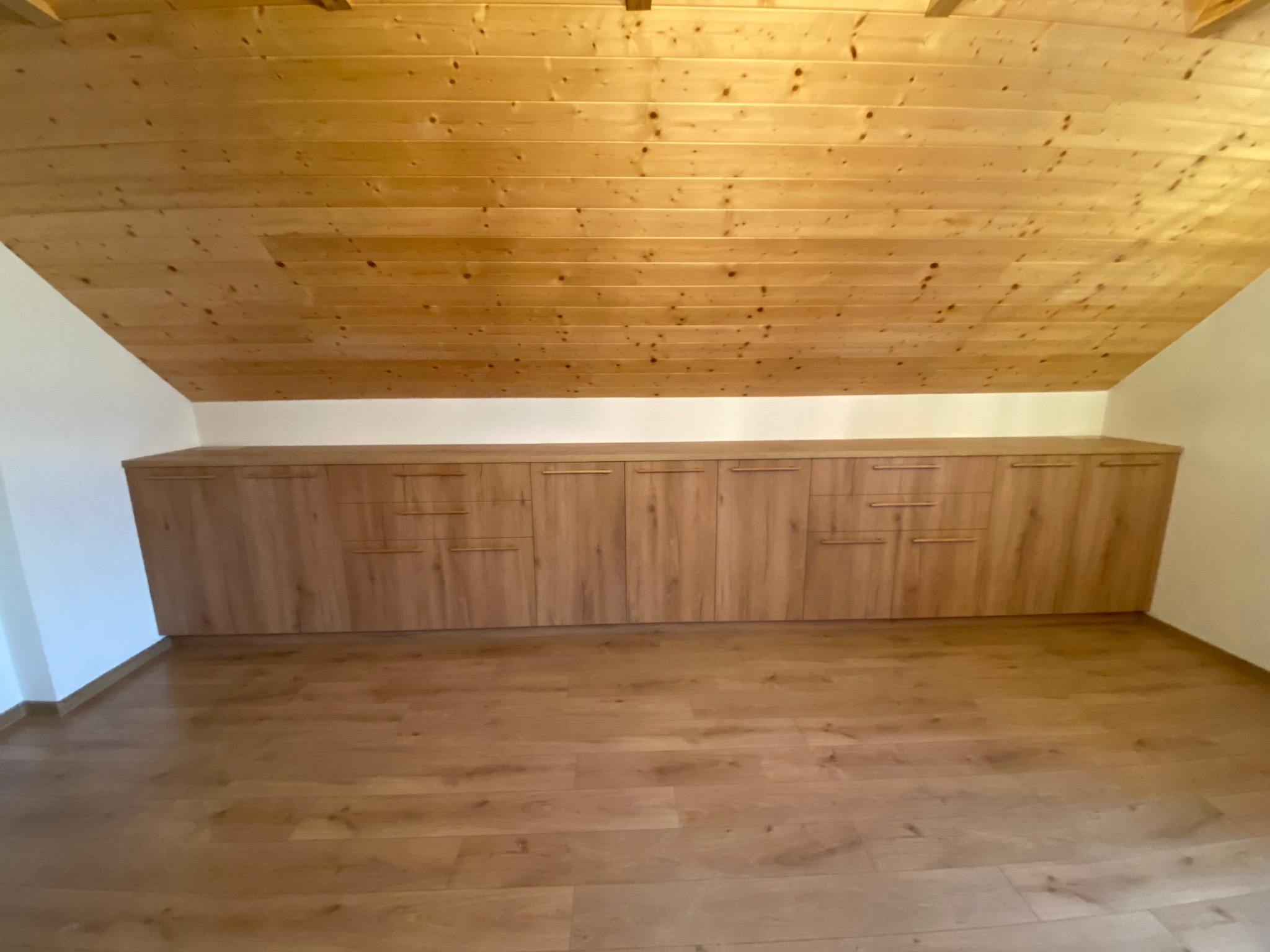 Szafa wnękowa z drewnianymi frontami, wkomponowana w skos dachu na poddaszu. Jasne drewno na podłodze i suficie tworzy spójną całość.