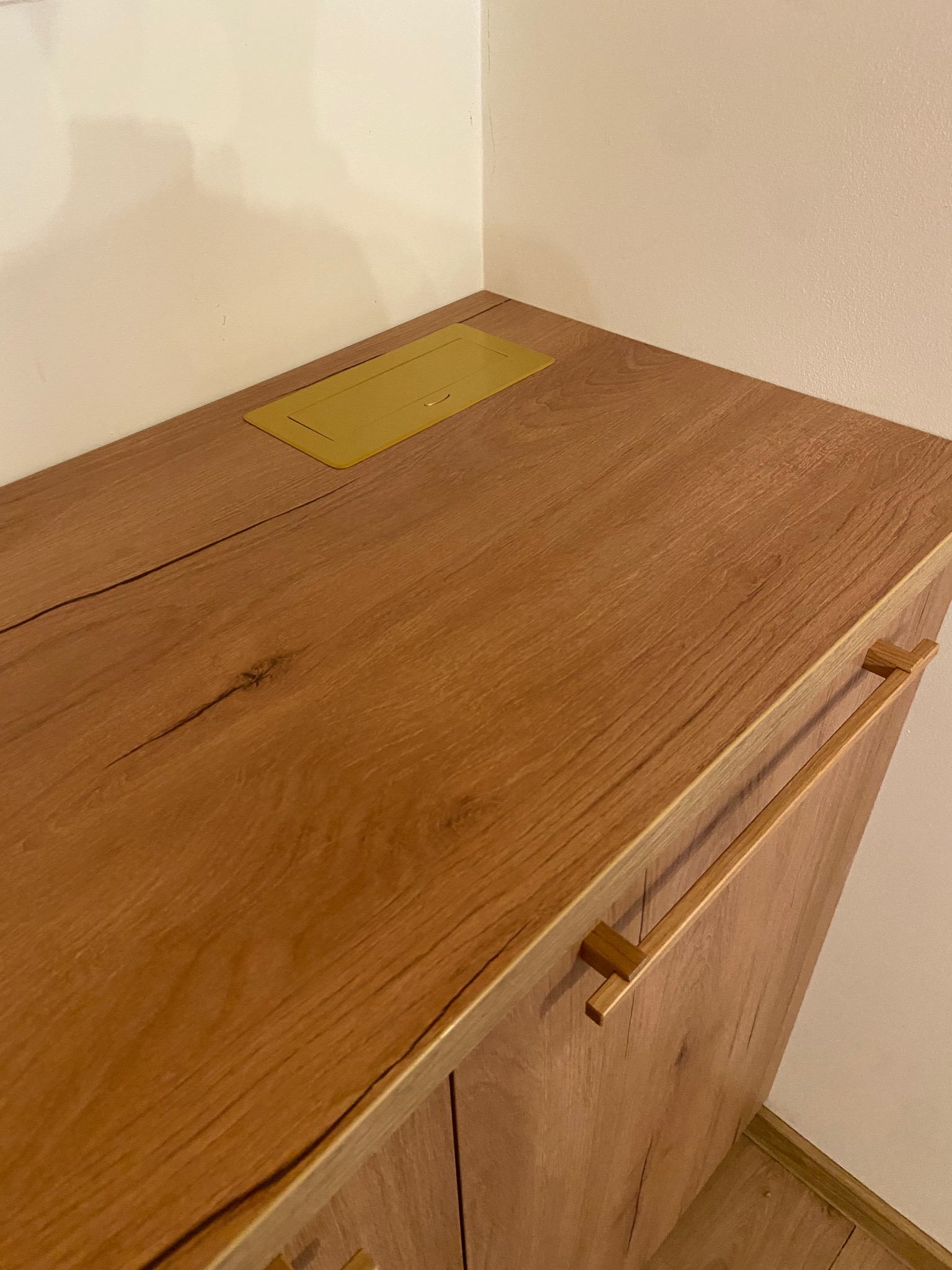 Fragment szafki z blatem o drewnianej fakturze i złotymi uchwytami. Na blacie widoczna złota klapka gniazda elektrycznego. Ujęcie z bliska, ukazujące detale wykończenia.