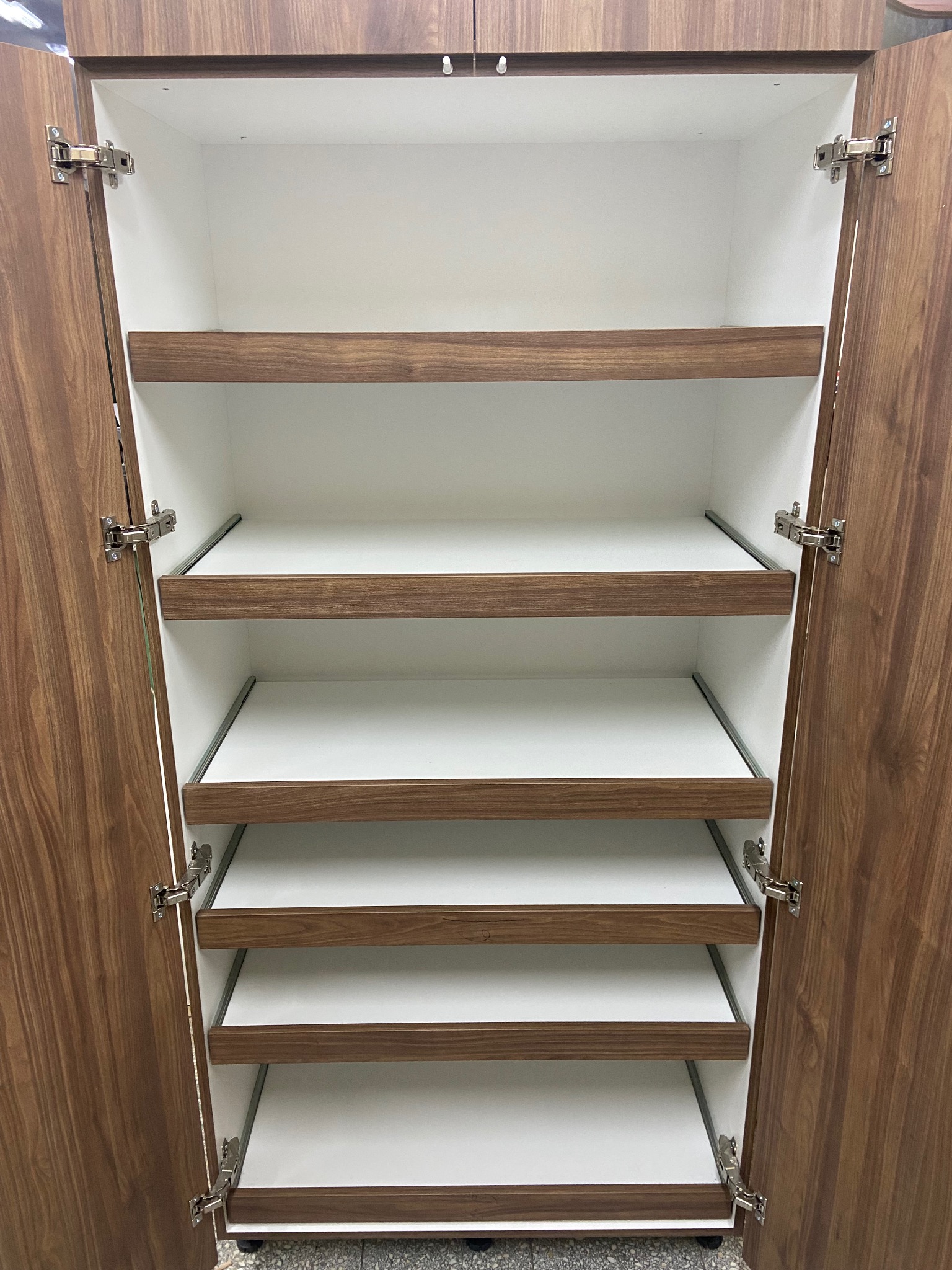 Otwarta szafa z półkami w kolorze drewna i białym wnętrzem. Widoczne zawiasy i metalowe prowadnice półek. Szafa z fornirem o wyraźnym rysunku słojów.