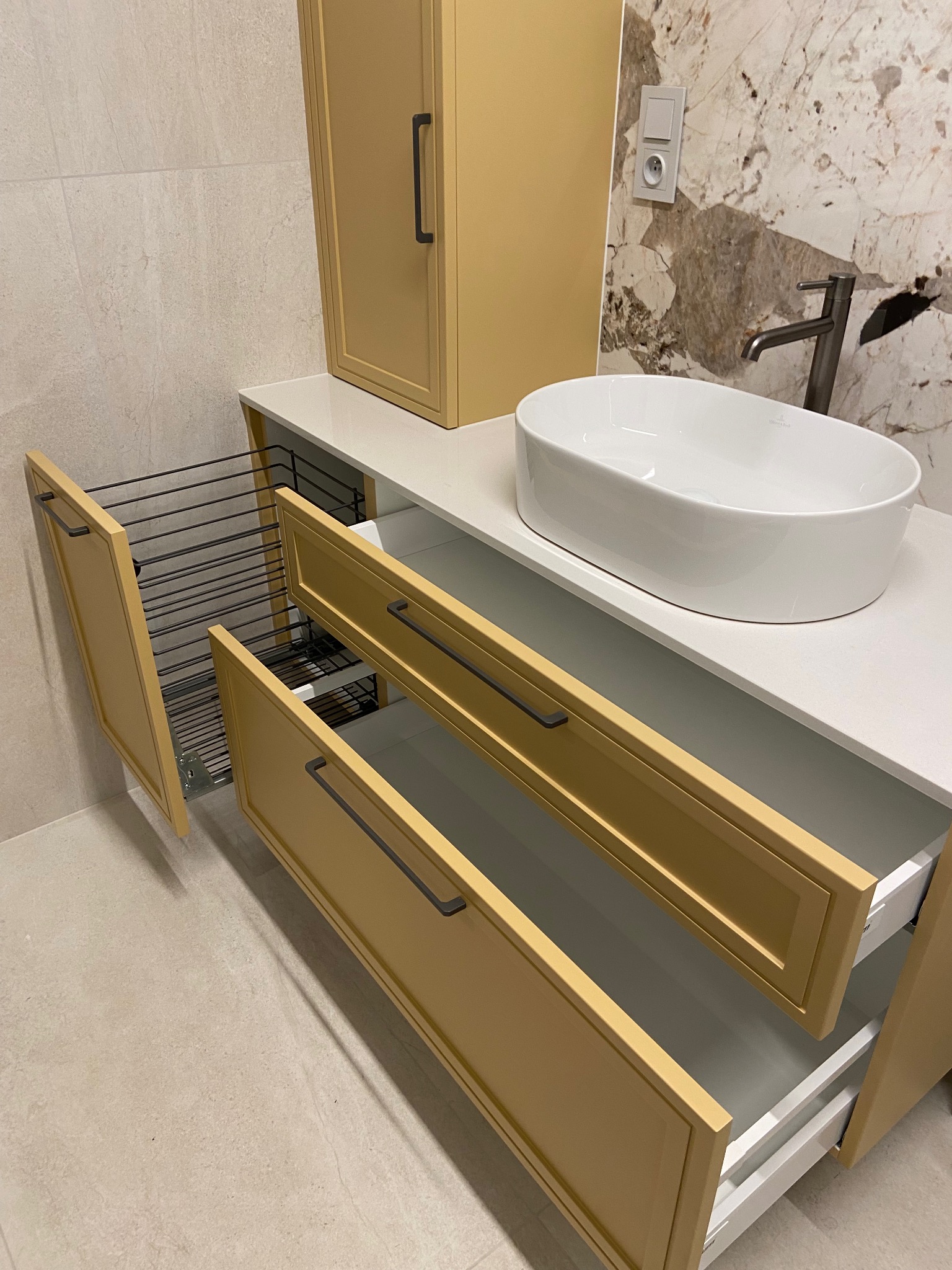 Żółta szafka łazienkowa z wysuwanymi szufladami i koszem na pranie, biała umywalka nablatowa, bateria, marmurowe wykończenie ściany w tle.