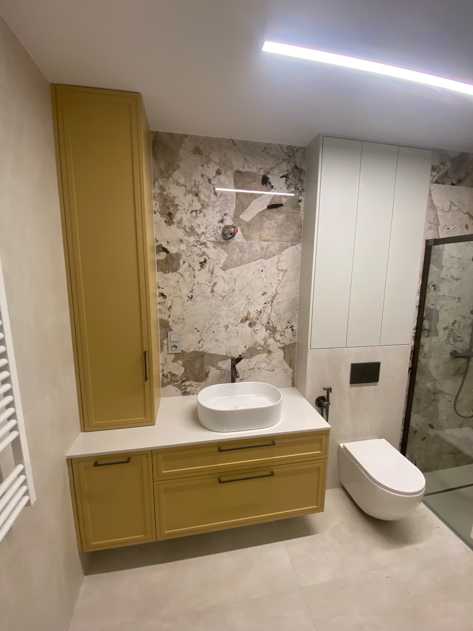 Nowoczesna łazienka z szafką pod umywalką w kolorze musztardowym, białą umywalką nablatową i marmurową ścianą. Minimalistyczny design, oświetlenie LED.