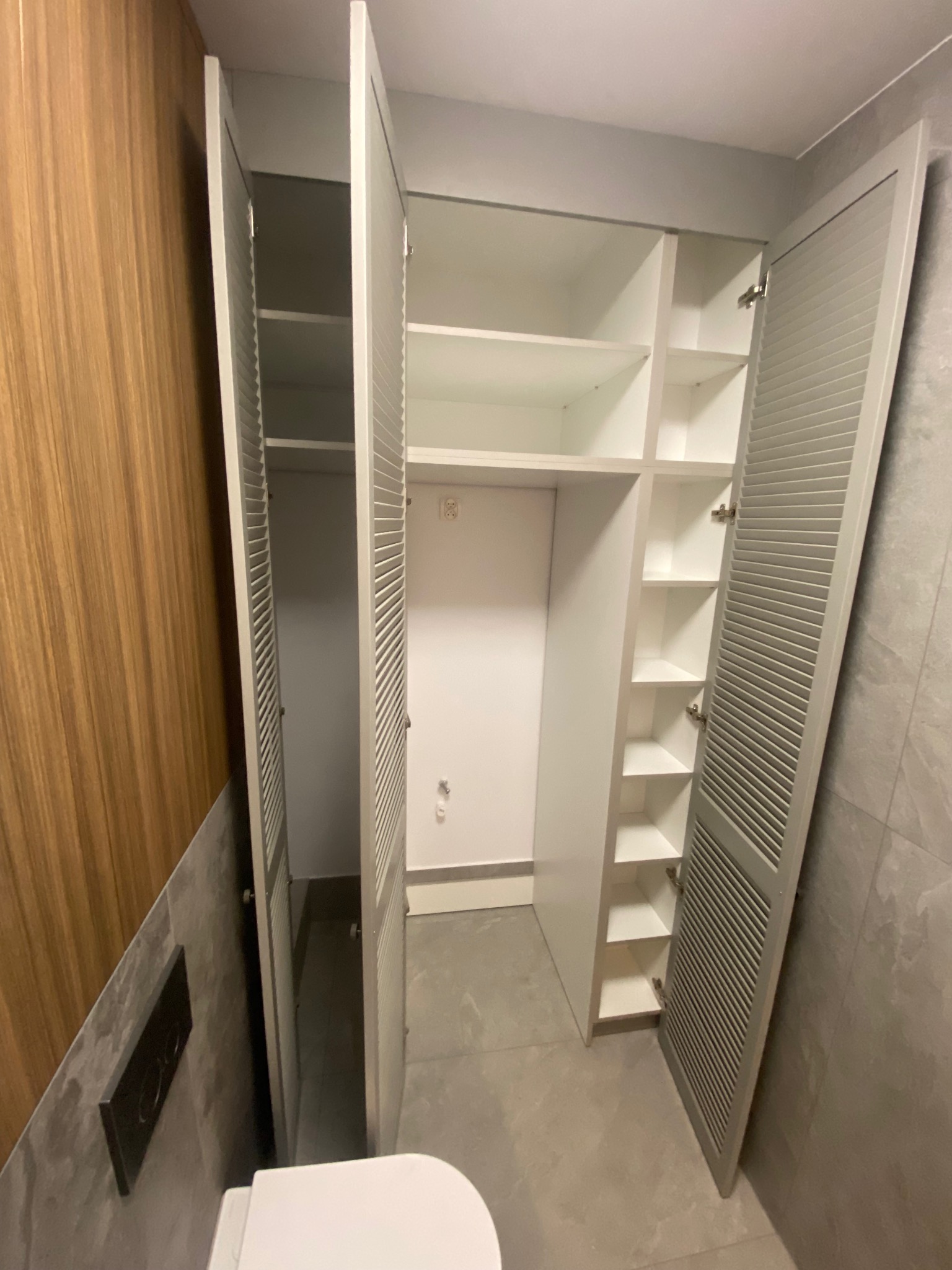 Biała szafa wnękowa z lamelowymi drzwiami, otwarte skrzydła ukazują półki. Widok z perspektywy toalety, szara płytka na ścianie i podłodze.