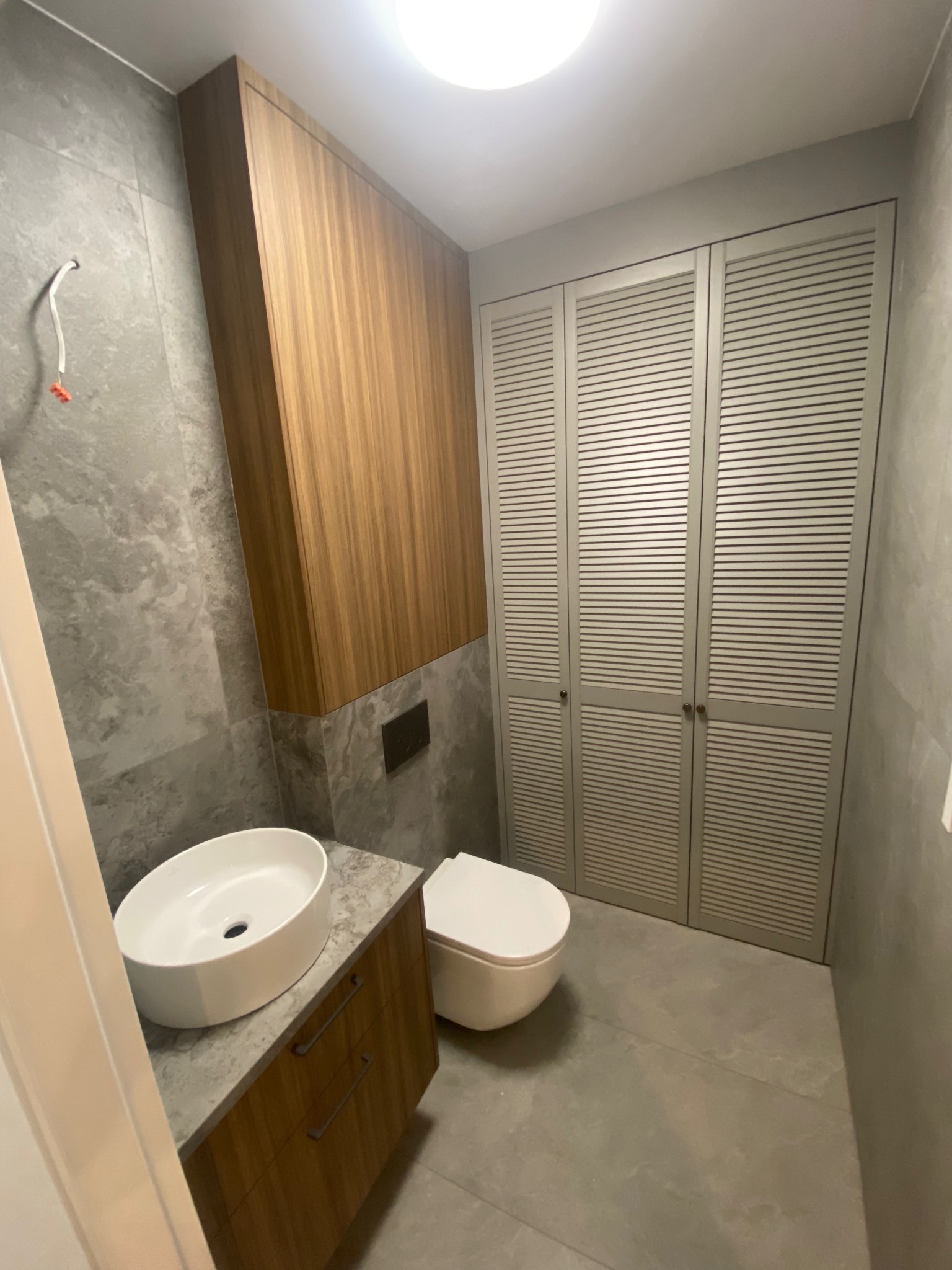 Nowoczesna łazienka z szafką pod umywalką z drewnianymi frontami, wiszącą szafką i szafą z lamelowymi drzwiami w szarym odcieniu.