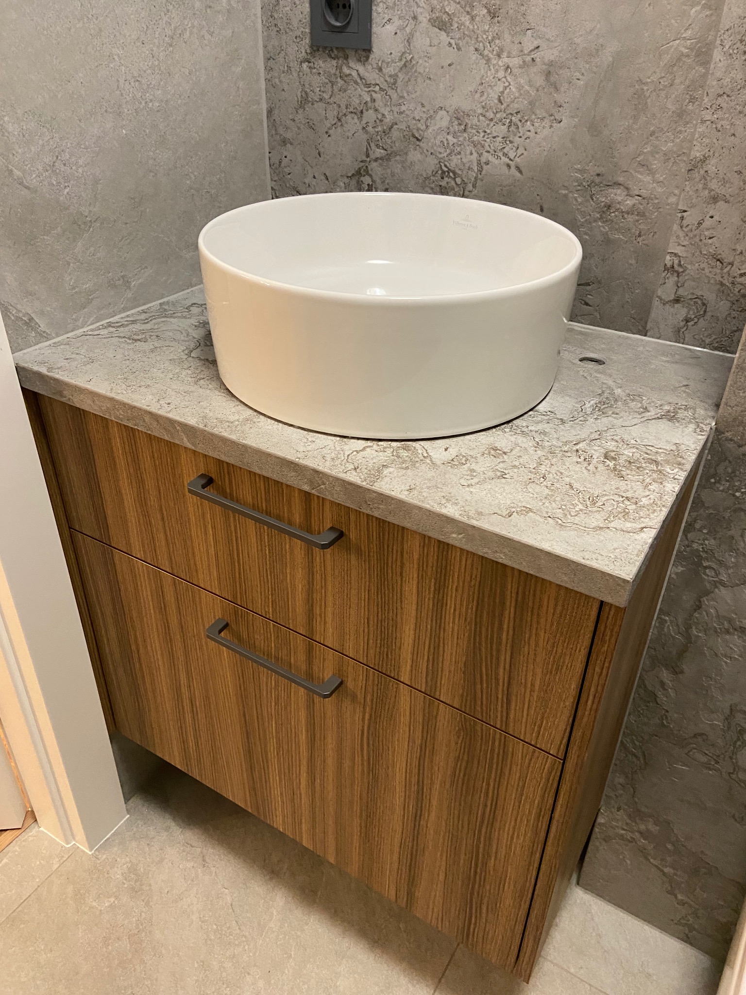 Nowoczesna szafka łazienkowa z drewnianymi frontami i umywalką nablatową Villeroy&Boch. Blat imitujący kamień, szare płytki w tle.