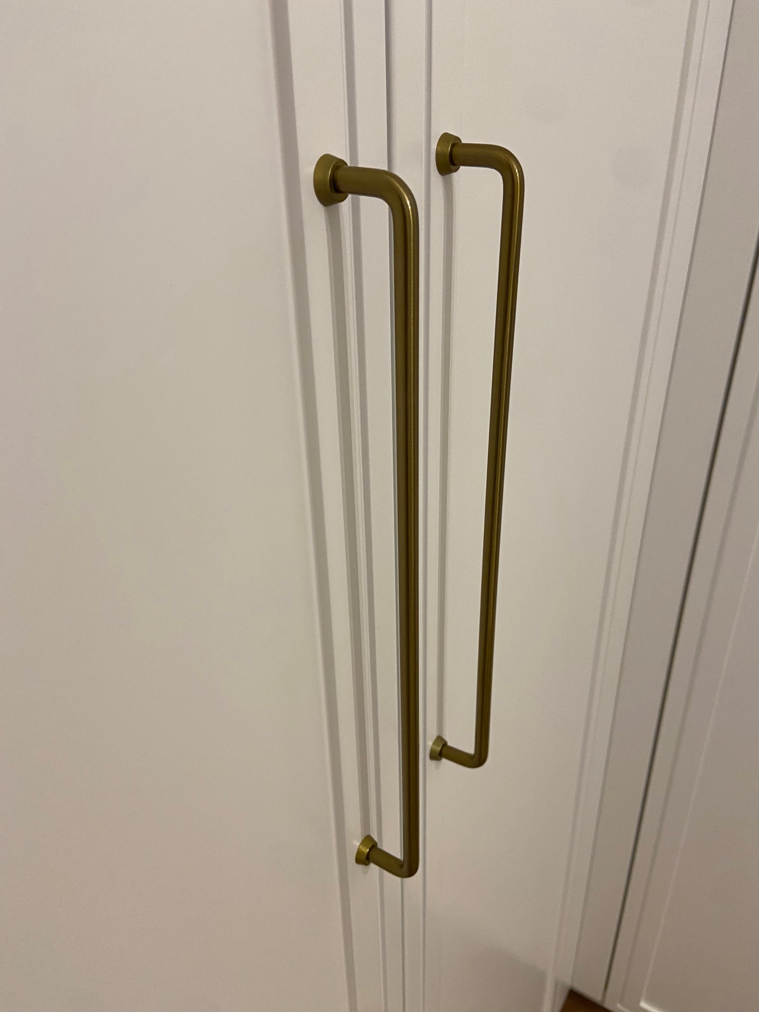 Białe drzwi szafy z długimi, złotymi uchwytami w kształcie litery L. Minimalistyczny design, subtelne oświetlenie.