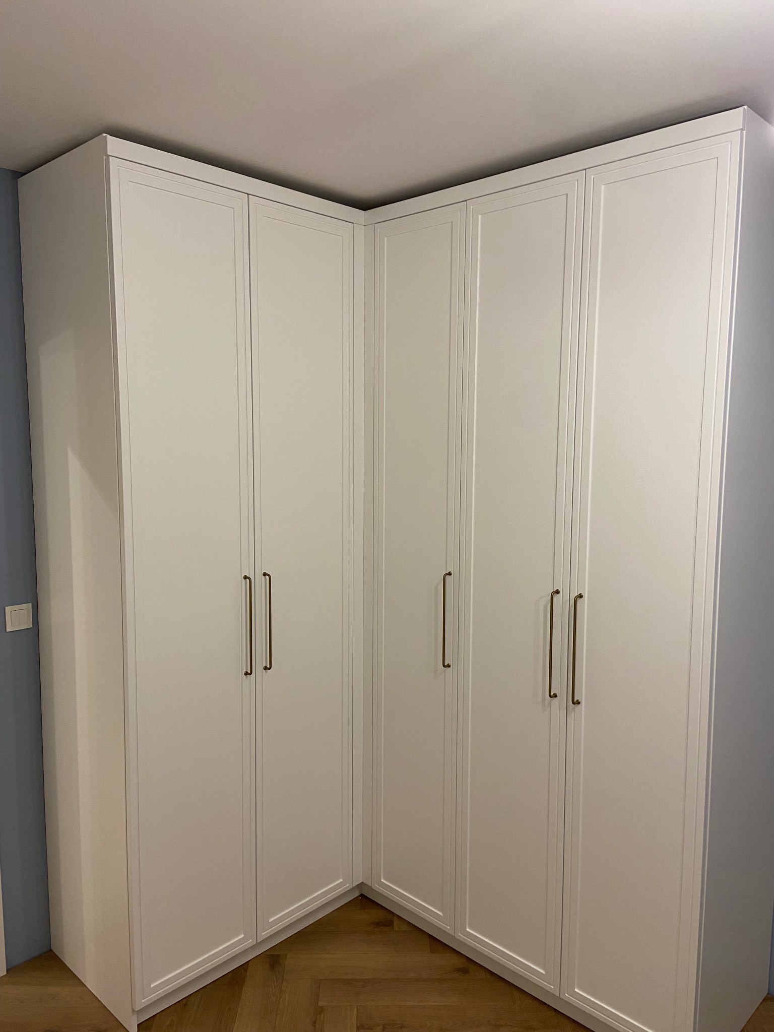 Biała, narożna szafa wnękowa z frezowanymi frontami i długimi, złotymi uchwytami, dopasowana do skosu sufitu, na tle błękitnej ściany i parkietu.