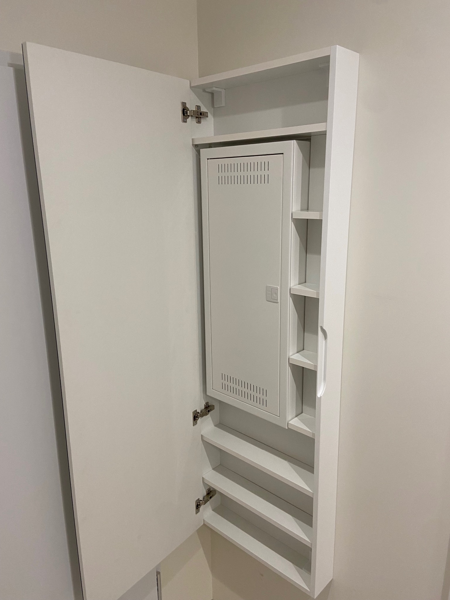 Biała szafa wnękowa z otwartymi drzwiami, ukazująca wewnętrzne półki i zabudowany panel elektryczny. Minimalistyczny design, jasne oświetlenie.