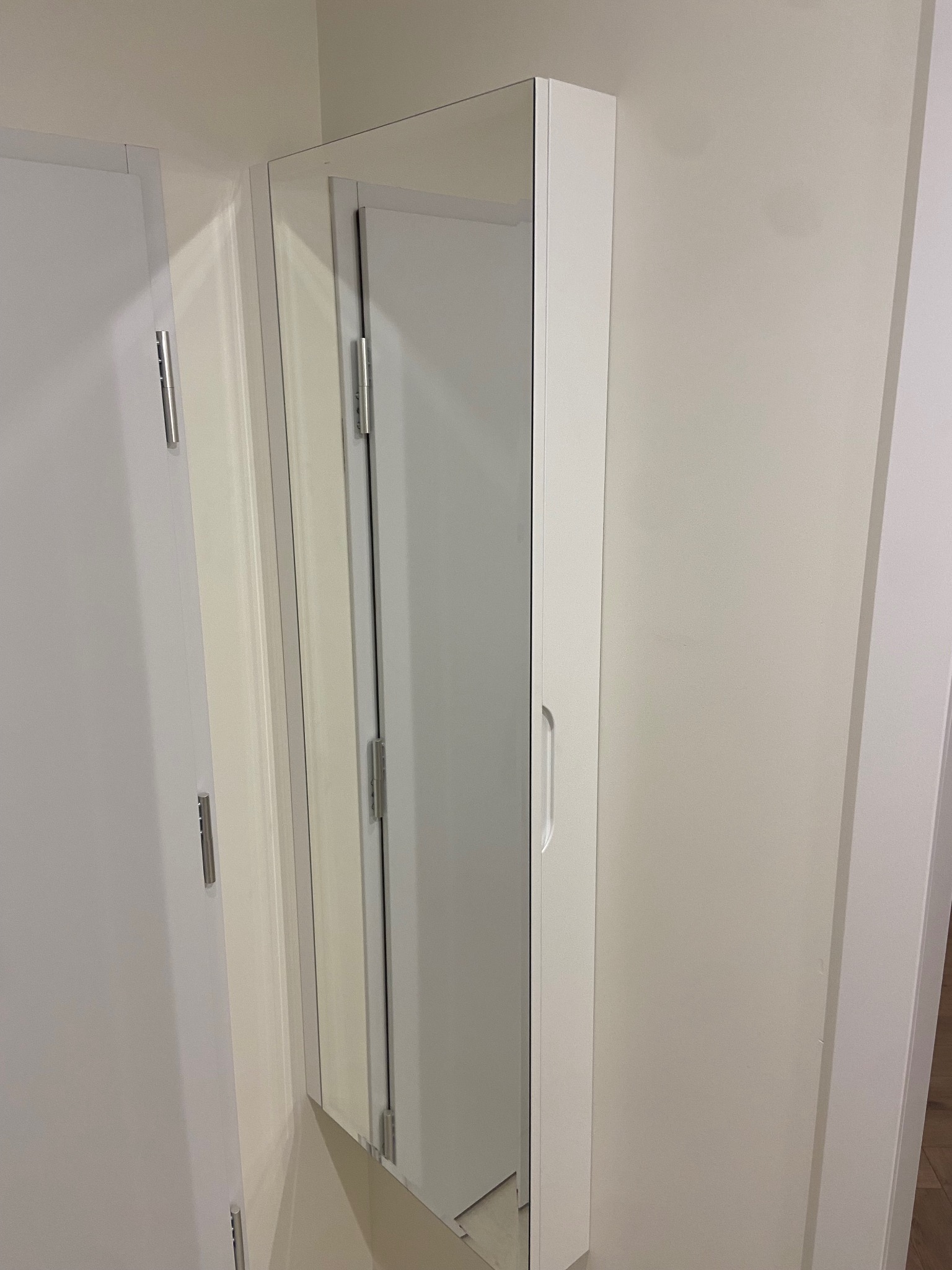 Biała szafka z lustrem, zamontowana na ścianie obok drzwi. Minimalistyczny design, widoczne zawiasy i uchwyt. Jasne wnętrze odbija się w lustrze.