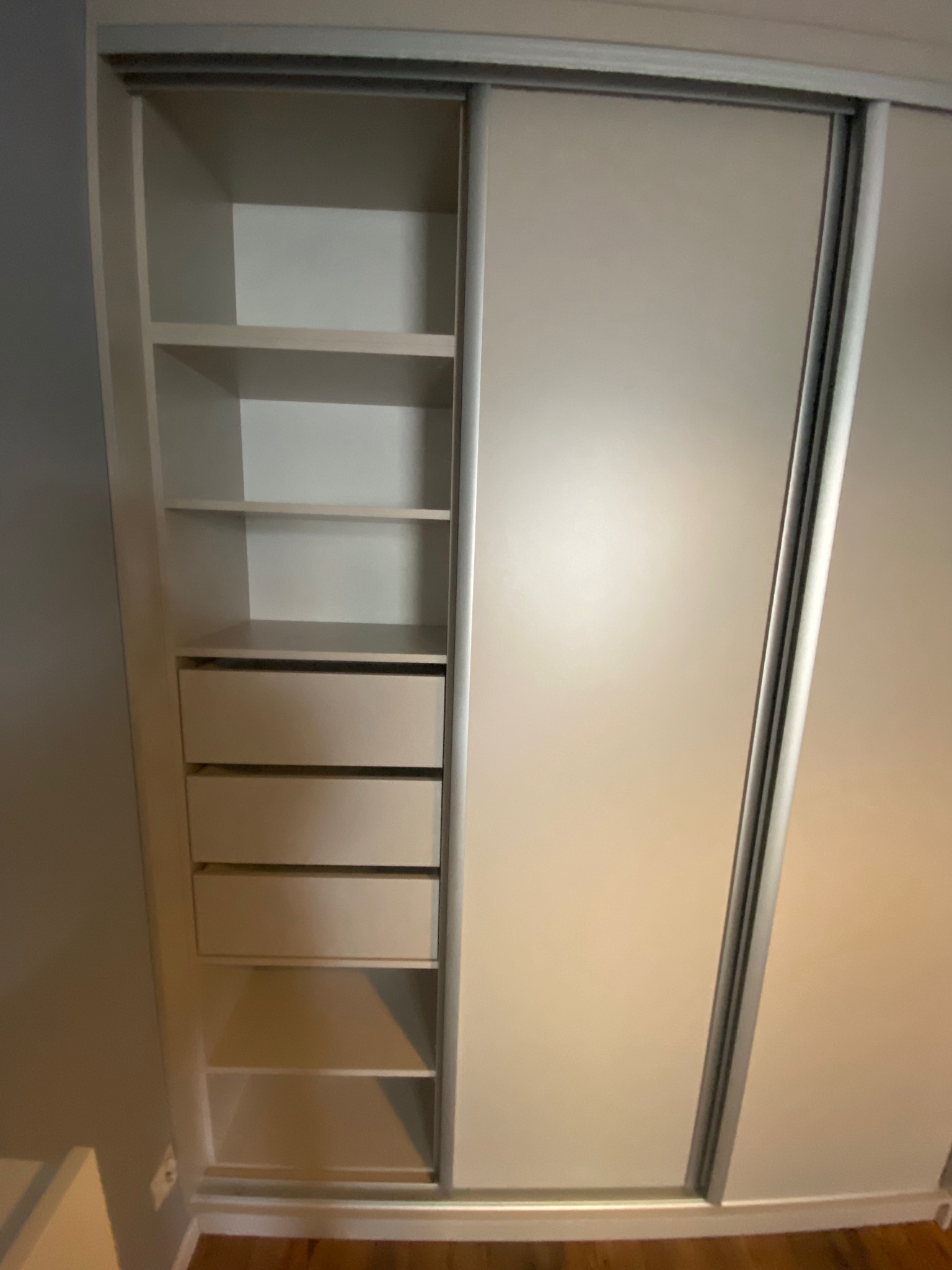 Jasna szafa wnękowa z przesuwnymi drzwiami i otwartymi półkami oraz szufladami, widok z bliska, srebrne obramowanie drzwi.