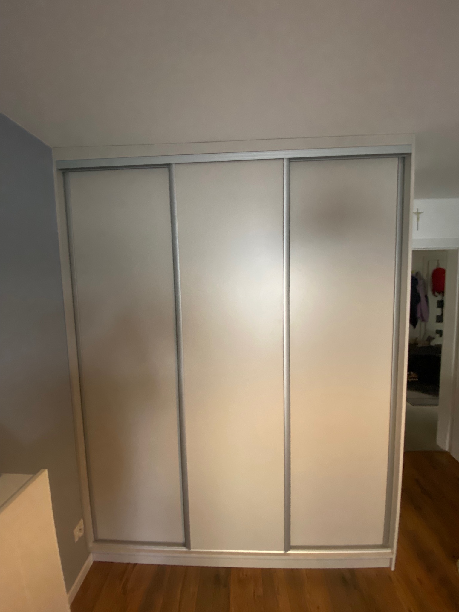 Jasna szafa przesuwna z trzema drzwiami, srebrne obramowanie, widok na wnętrze pokoju z krzyżem na ścianie. Parkiet na podłodze.