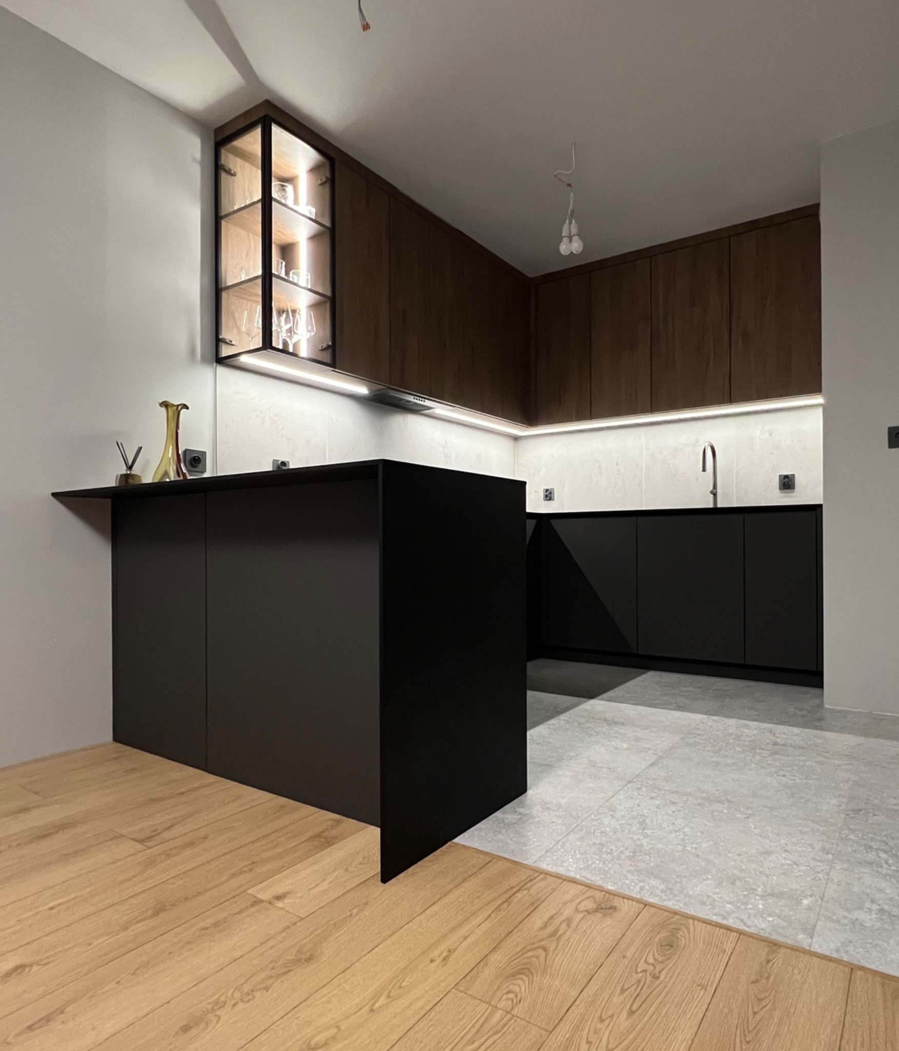 Nowoczesna kuchnia z czarną wyspą i drewnianymi szafkami. Podświetlane półki z kieliszkami. Kontrastujące podłogi: drewniana i szara płytka. Minimalistyczny design.