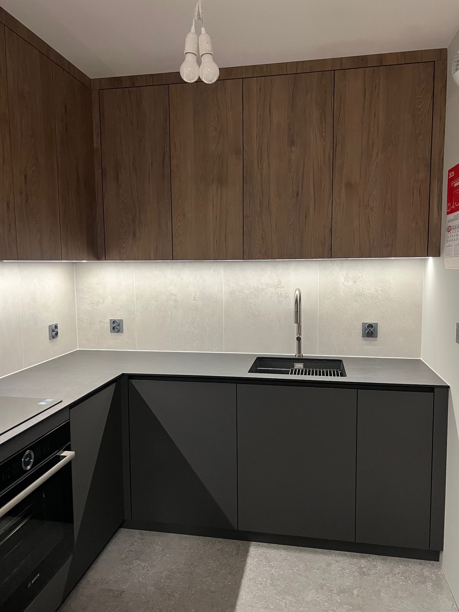 Nowoczesna kuchnia z ciemnymi szafkami dolnymi i drewnopodobnymi górnymi, oświetlona listwą LED. Minimalistyczny design z czarnym zlewem i baterią.