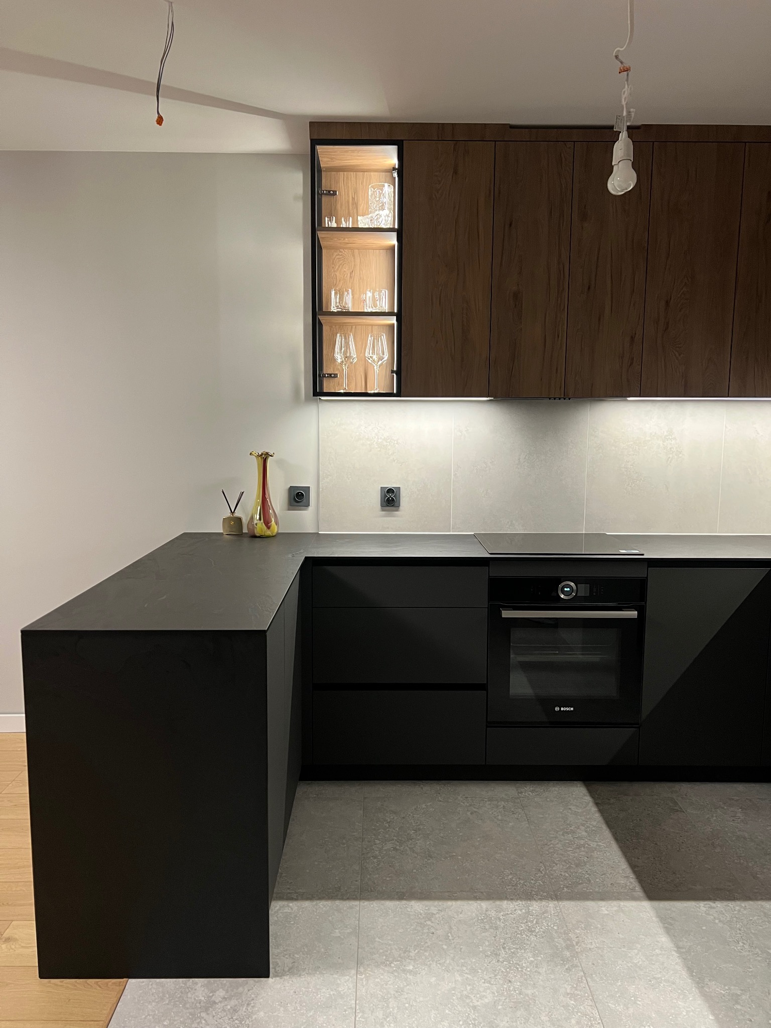 Nowoczesna kuchnia: czarne matowe fronty, drewniane szafki wiszące, szklana witryna z oświetleniem. Minimalistyczny design, szary blat i płytki. Wazon jako akcent kolorystyczny.