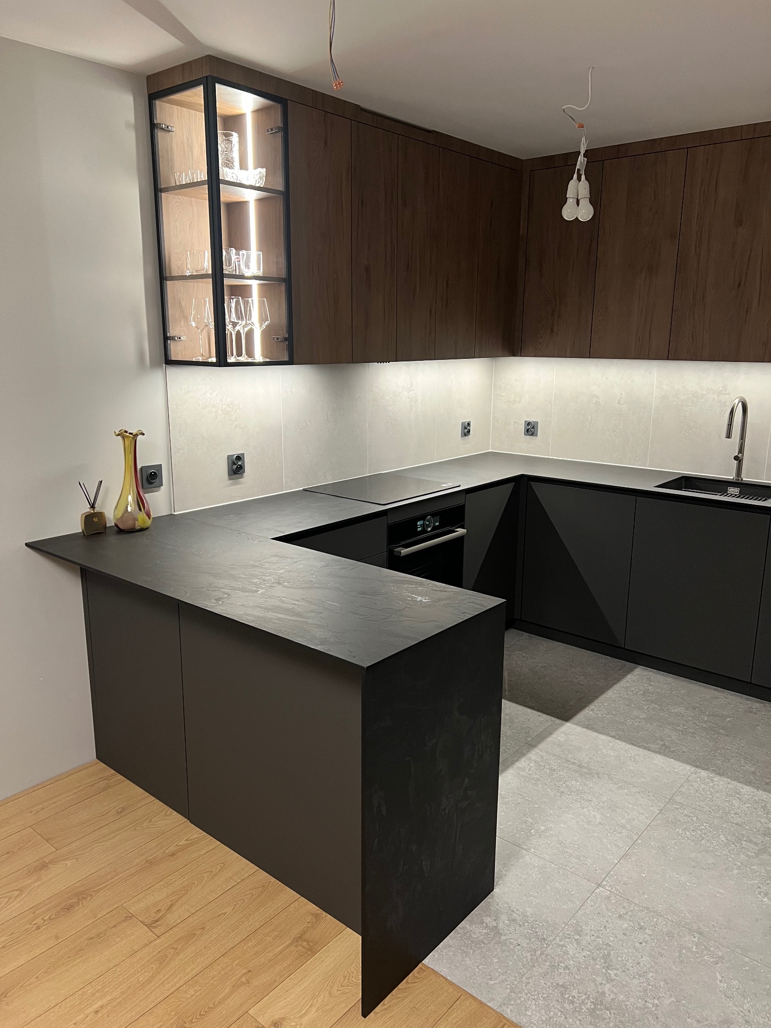 Nowoczesna kuchnia w kształcie litery U z ciemnymi frontami, blatem imitującym kamień i przeszkloną witryną z podświetleniem. Widoczne naczynia i dekoracje.