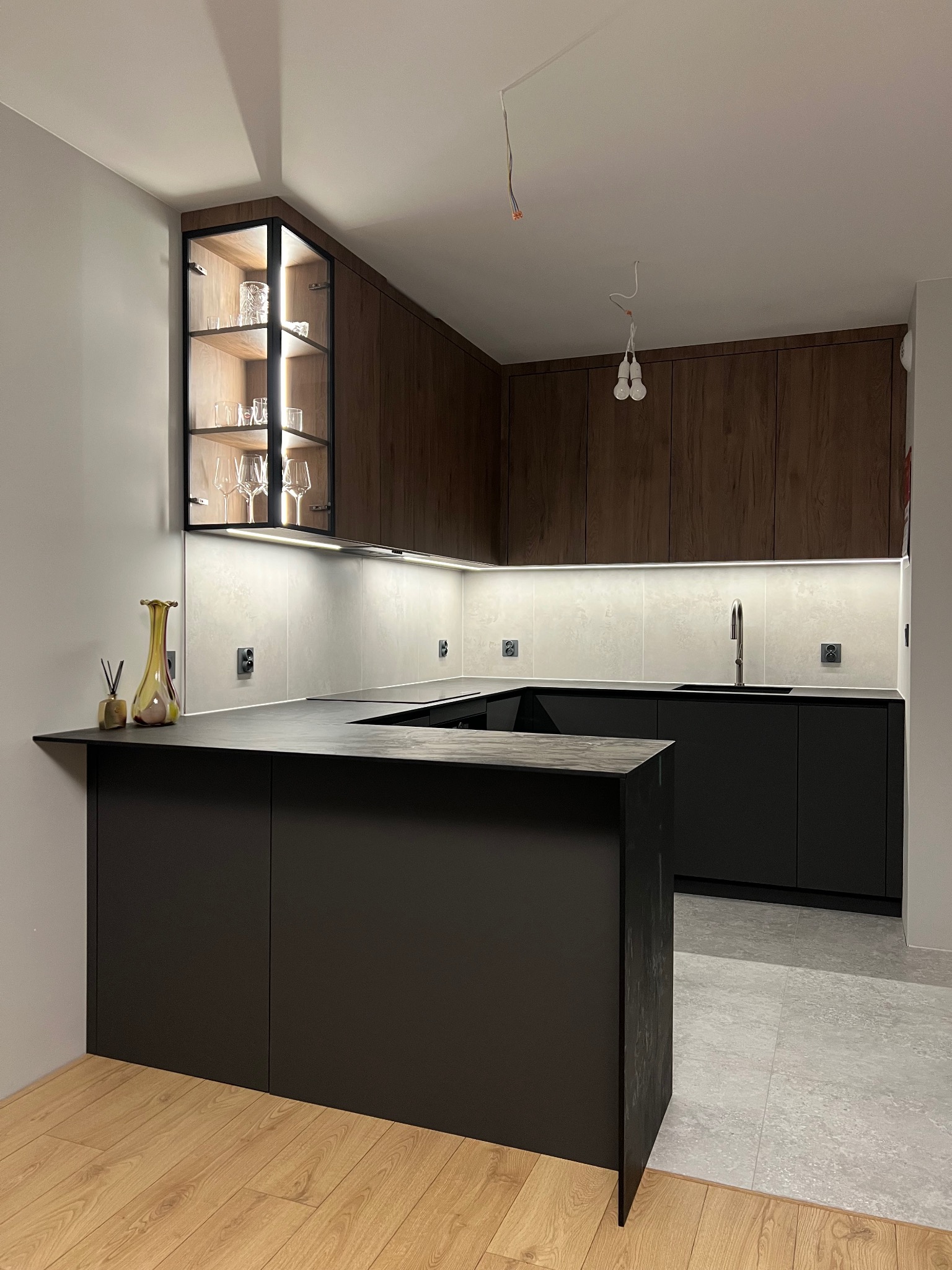 Nowoczesna kuchnia z ciemnymi frontami i drewnianymi akcentami. Podświetlane szafki wiszące z ekspozycją szkła. Minimalistyczny design z wyspą kuchenną i oświetleniem LED.