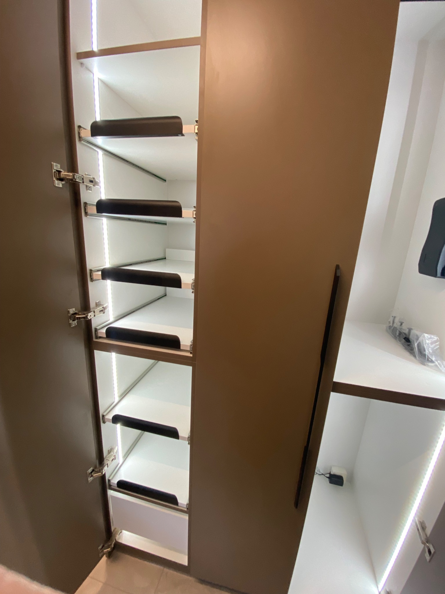 Otwarta szafa wnękowa z oświetleniem LED, brązowe fronty, wysuwane półki z czarnymi uchwytami. Widoczne zawiasy i fragment wnętrza drugiej części szafy.