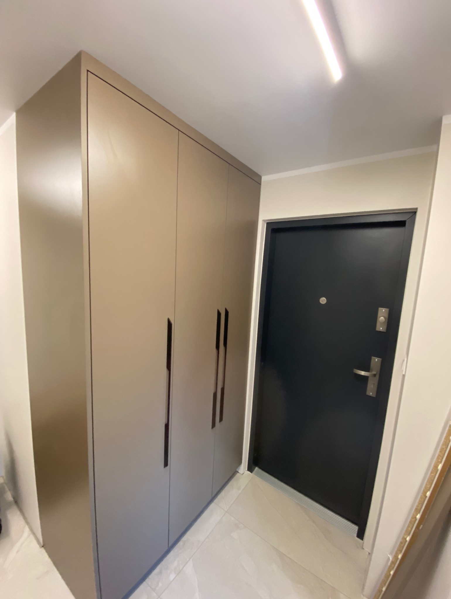 Beżowa szafa wnękowa z długimi, pionowymi uchwytami, umieszczona w przedpokoju obok czarnych drzwi wejściowych. Minimalistyczny design, jasne płytki podłogowe.