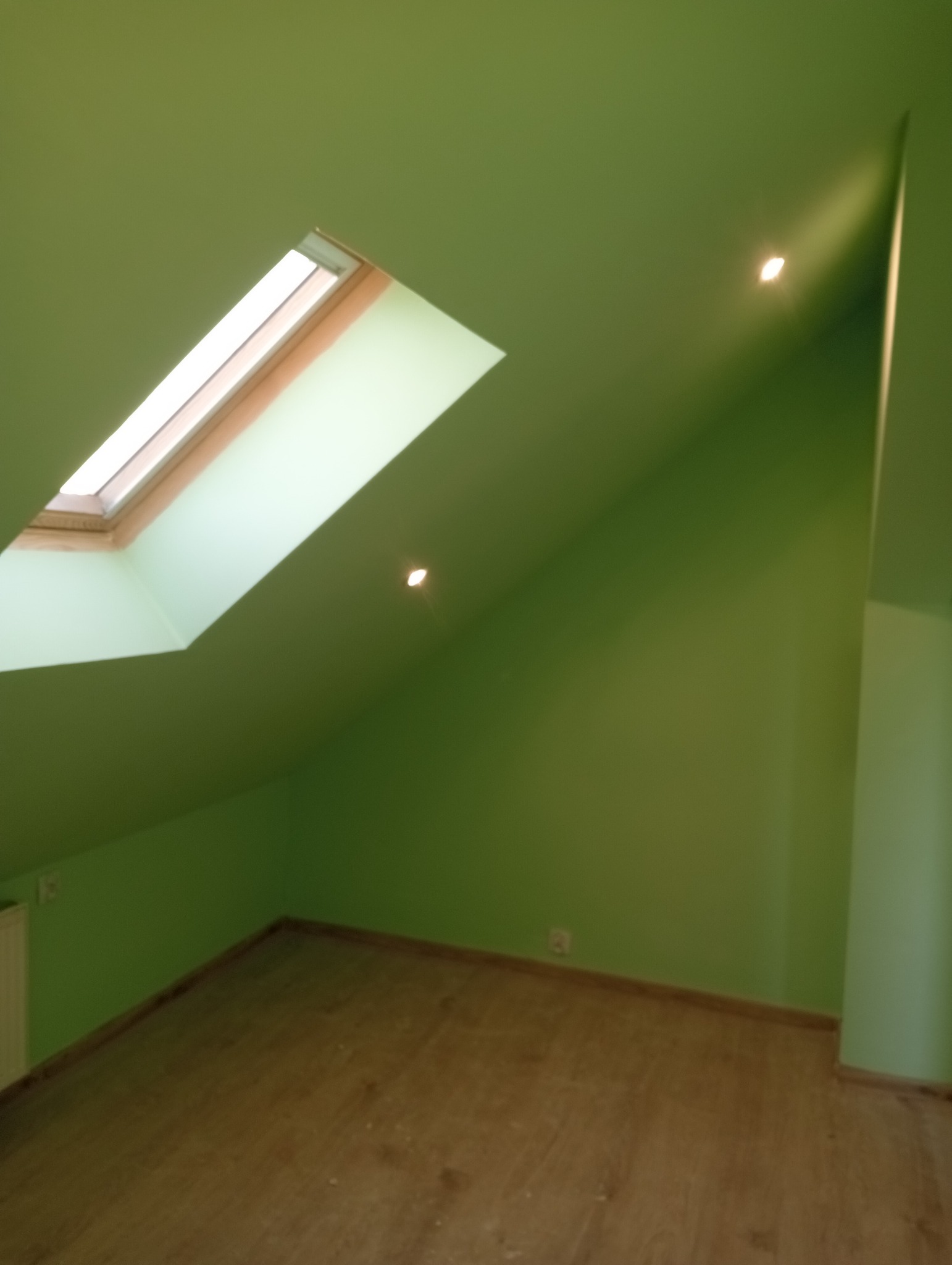 Poddasze po malowaniu na zielono z oknem dachowym i panelami podłogowymi, widoczne punkty świetlne w suficie.