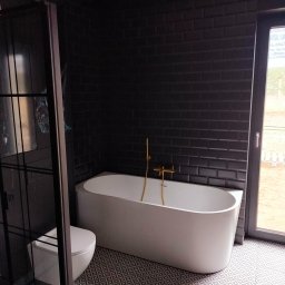 Art-dom - Nowoczesna łazienka z czarnymi płytkami typu metro, białą wanną z złotą armaturą, toaletą podwieszaną i geometryczną podłogą.