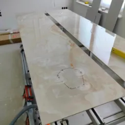 Przygotowanie blatu z płyty imitującej marmur z wyciętymi otworami na zlew, na stole do cięcia płytek, w tle okno i fragment ściany z dekoracyjną sztukaterią.