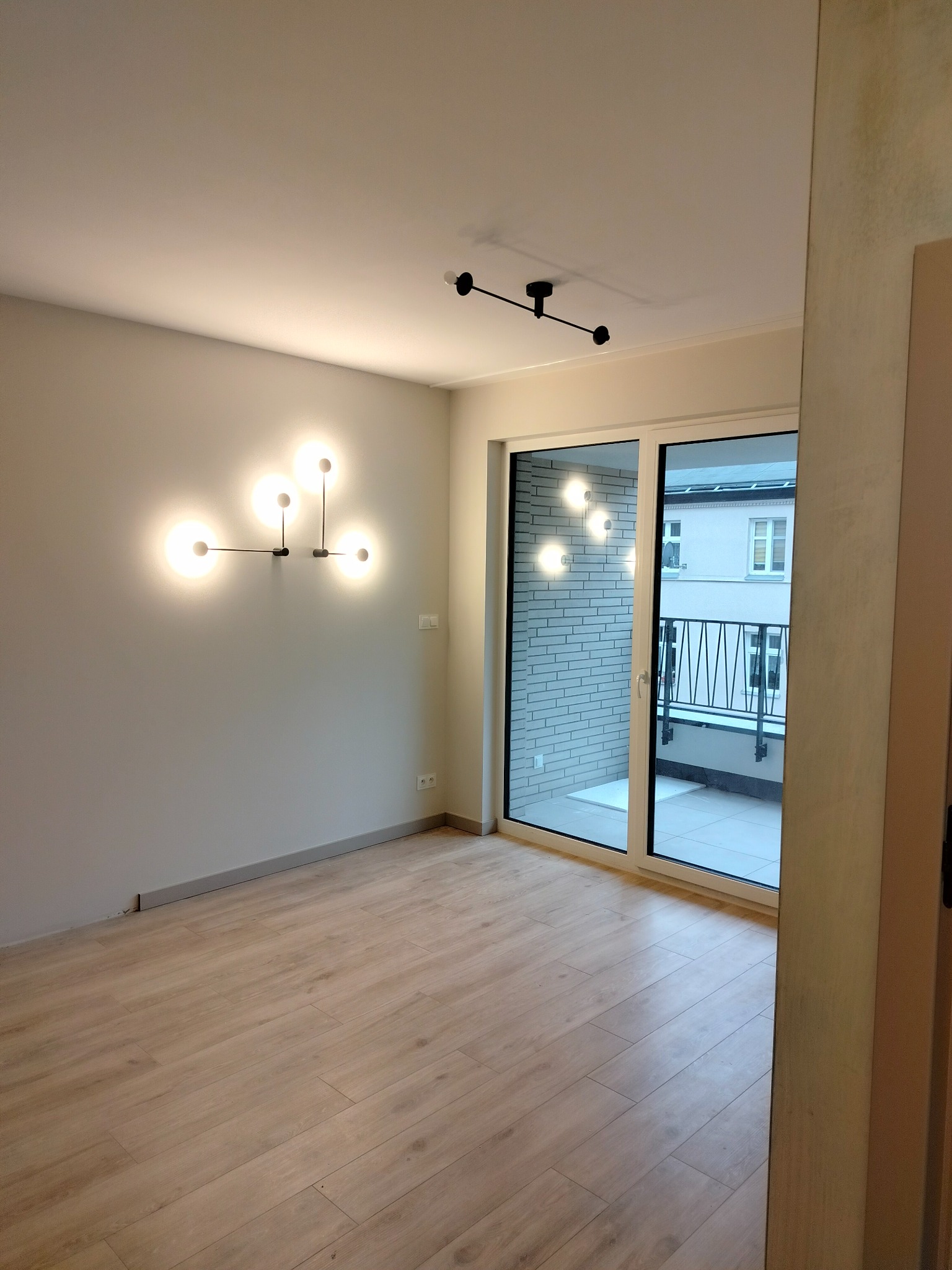 Jasne panele podłogowe w pustym pokoju z nowoczesnym oświetleniem ściennym i sufitowym, widok na balkon przez przesuwne drzwi balkonowe.